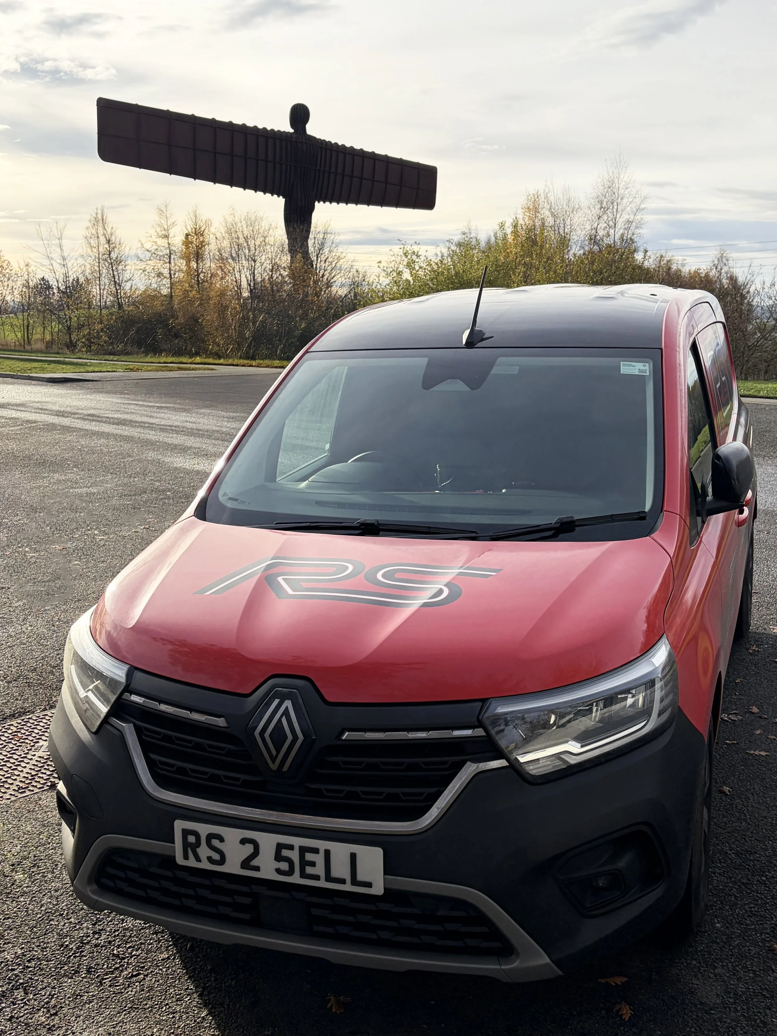 Red Renault car with RS logo on the hood, license plate RS 2 SELL, parked on a paved surface, with the Angel of the North in the background and trees with autumn foliage surrounding it.