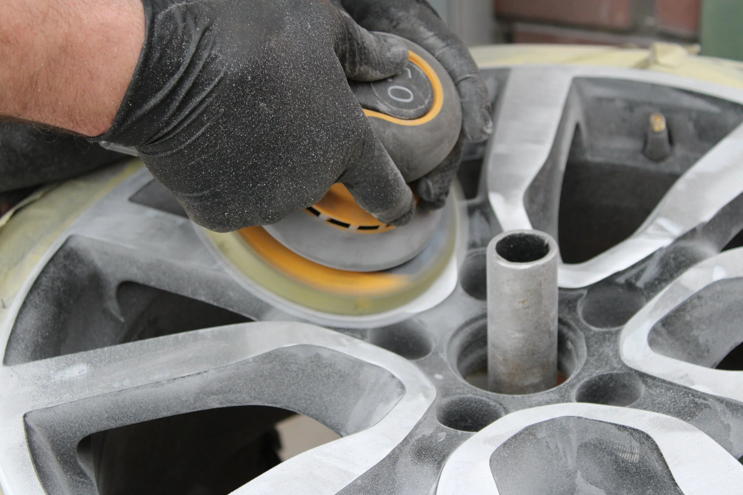A person wearing black gloves uses a handheld power tool to sand the surface of an alloy in preparation for repairs and refinishing.
