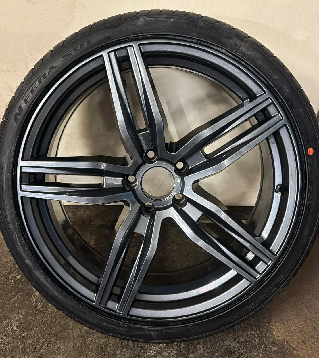 A close-up of an M3 Gunmetal, multi-spoke car wheel rim with intricate design, hanging on a rack with a blurred workshop background
