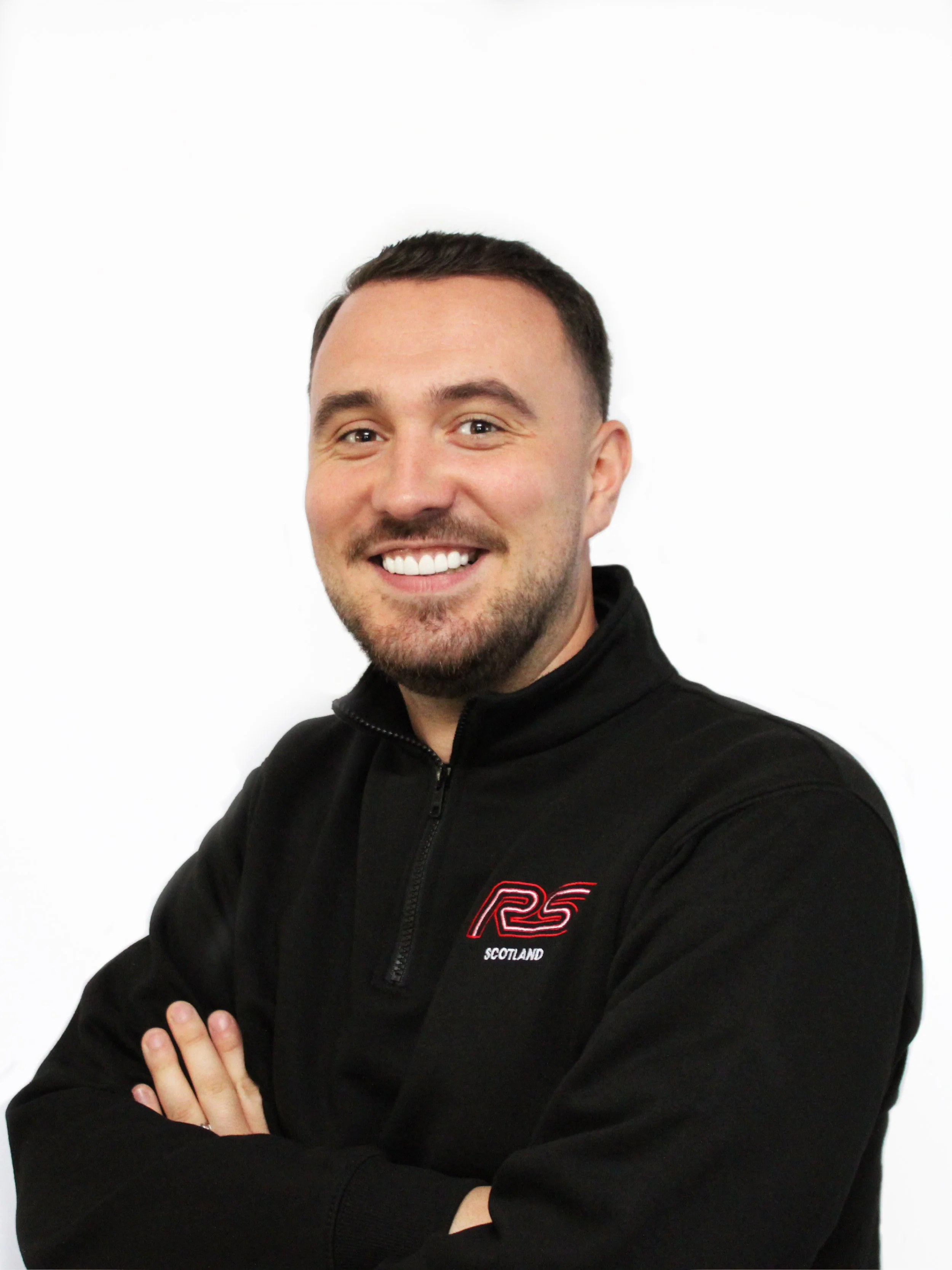 A man with short dark hair and a beard, smiling with crossed arms, wearing a black jacket with a logo on the chest that says 'RS Scotland' against a plain white background.