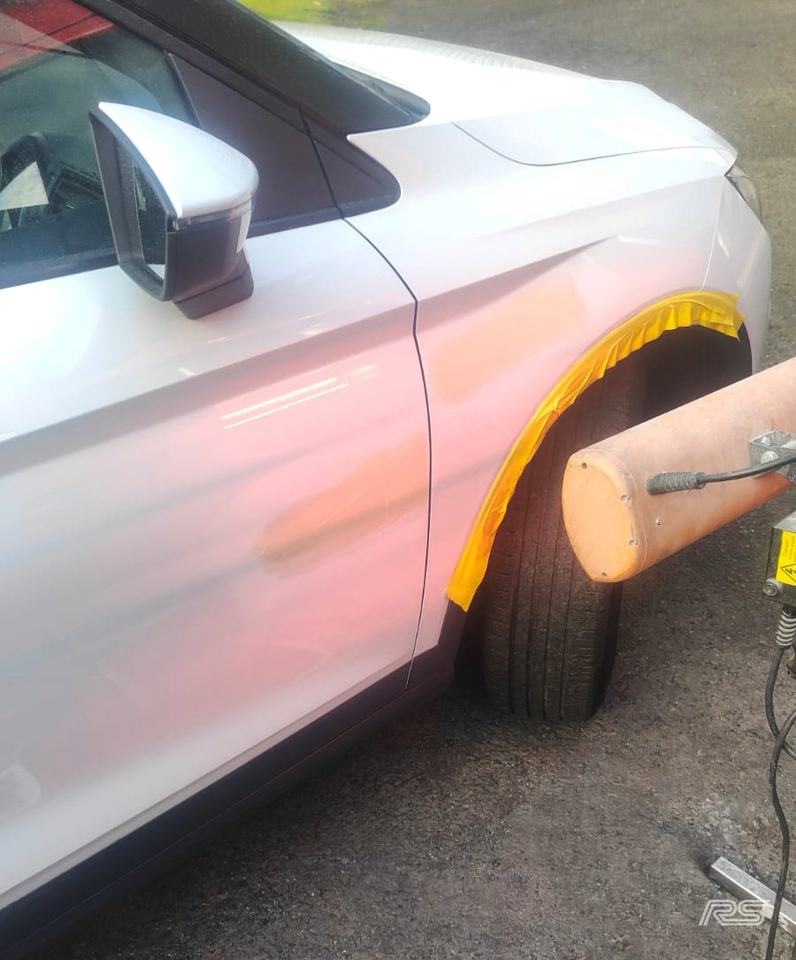 A white car being repaired or painted, with the wheel covered with yellow tape and a large heat lamp nearby for paint curing.