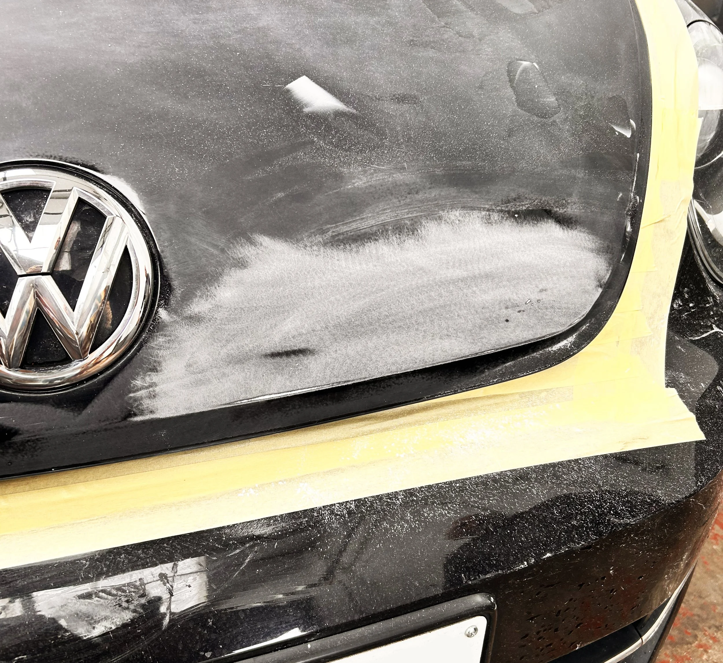 Close-up of a black Volkswagen car with visible scratches and paint damage on the front hood being prepared for repair, with masking tape around the edges.