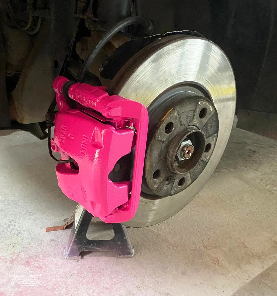 A car's brake rotor and pink brake caliper with a jack stand underneath.