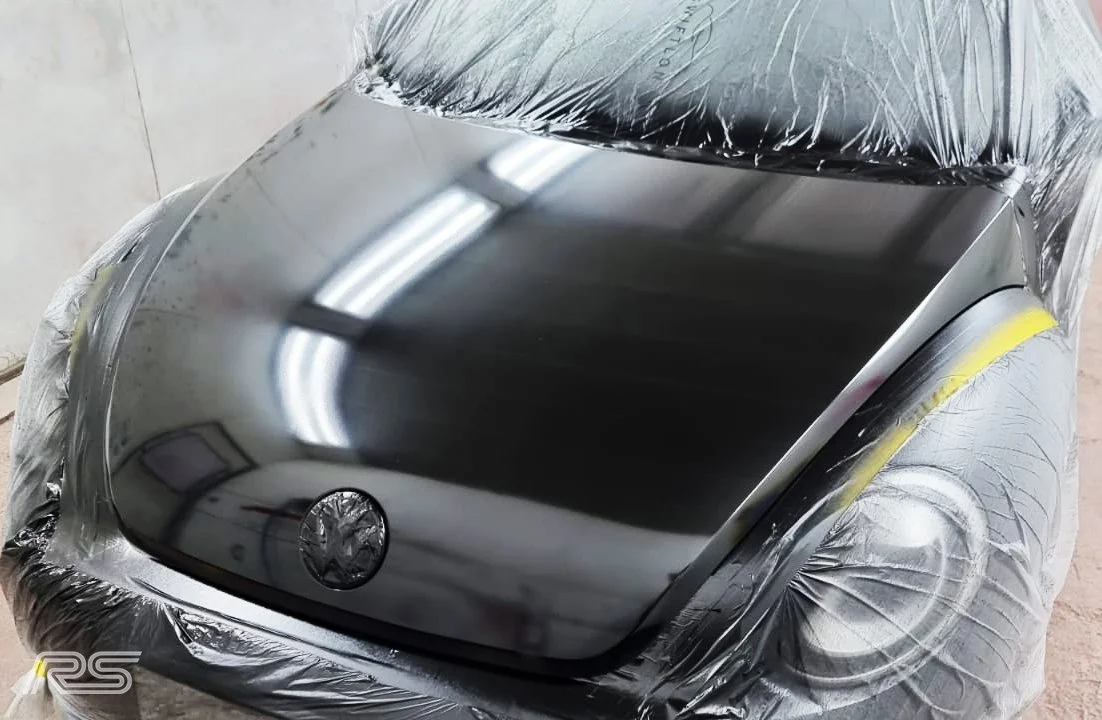 A shiny black Volkswagen car hood with the rest of the vehicle covered with plastic wrap.