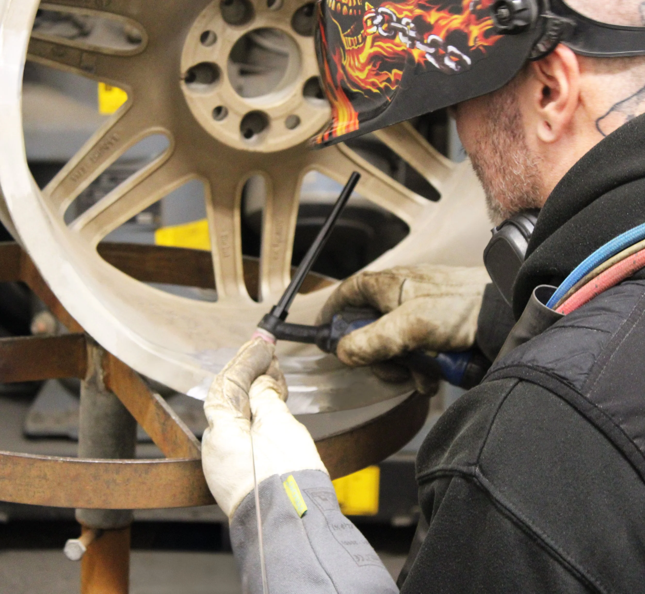 A person wearing a helmet and gloves is welding a large wheel with multiple spokes at the RS Wheels workshop.