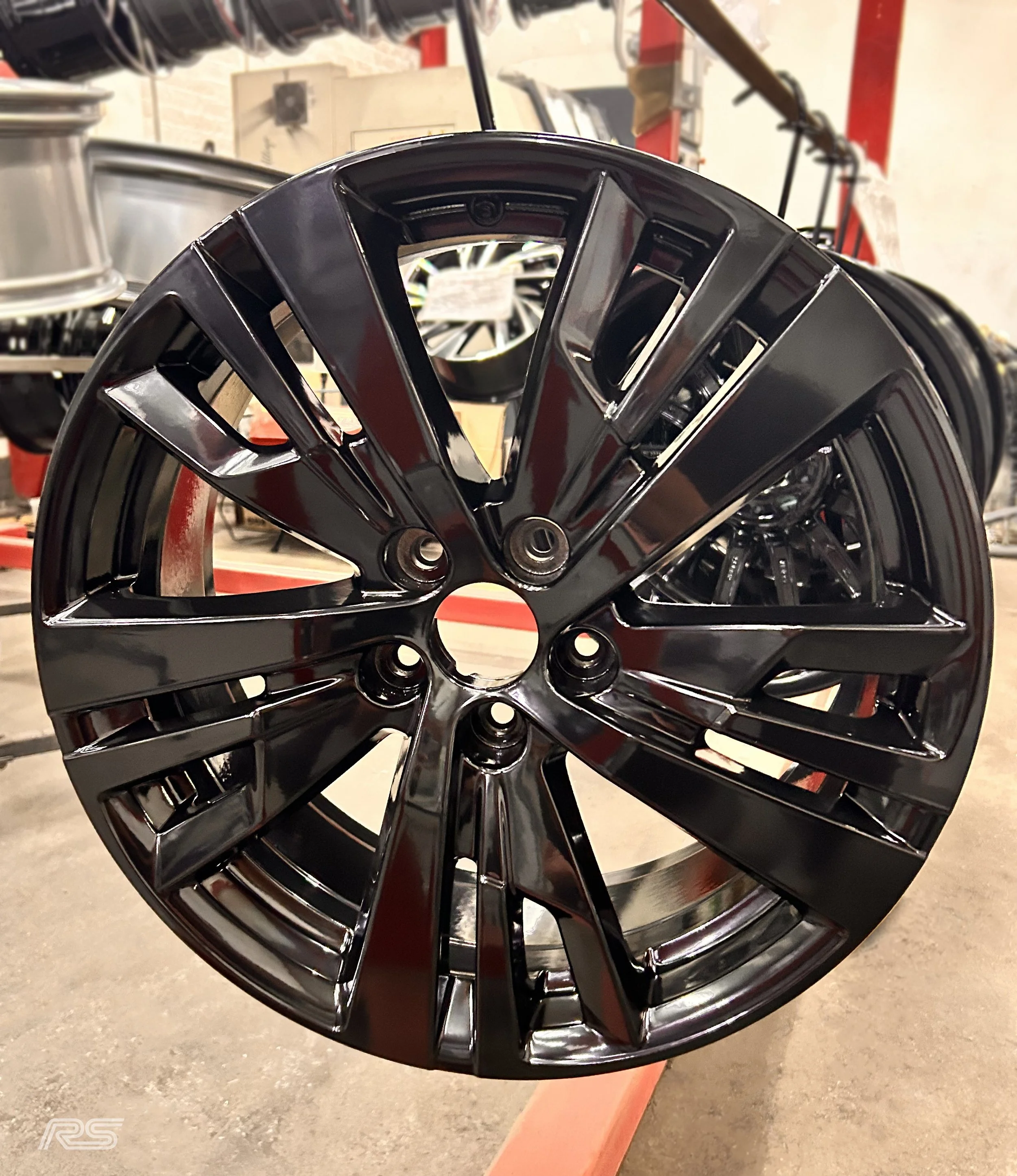 A black alloy  with multiple spokes, mounted on a hook in a workshop area during a powder coat refurbishment.
