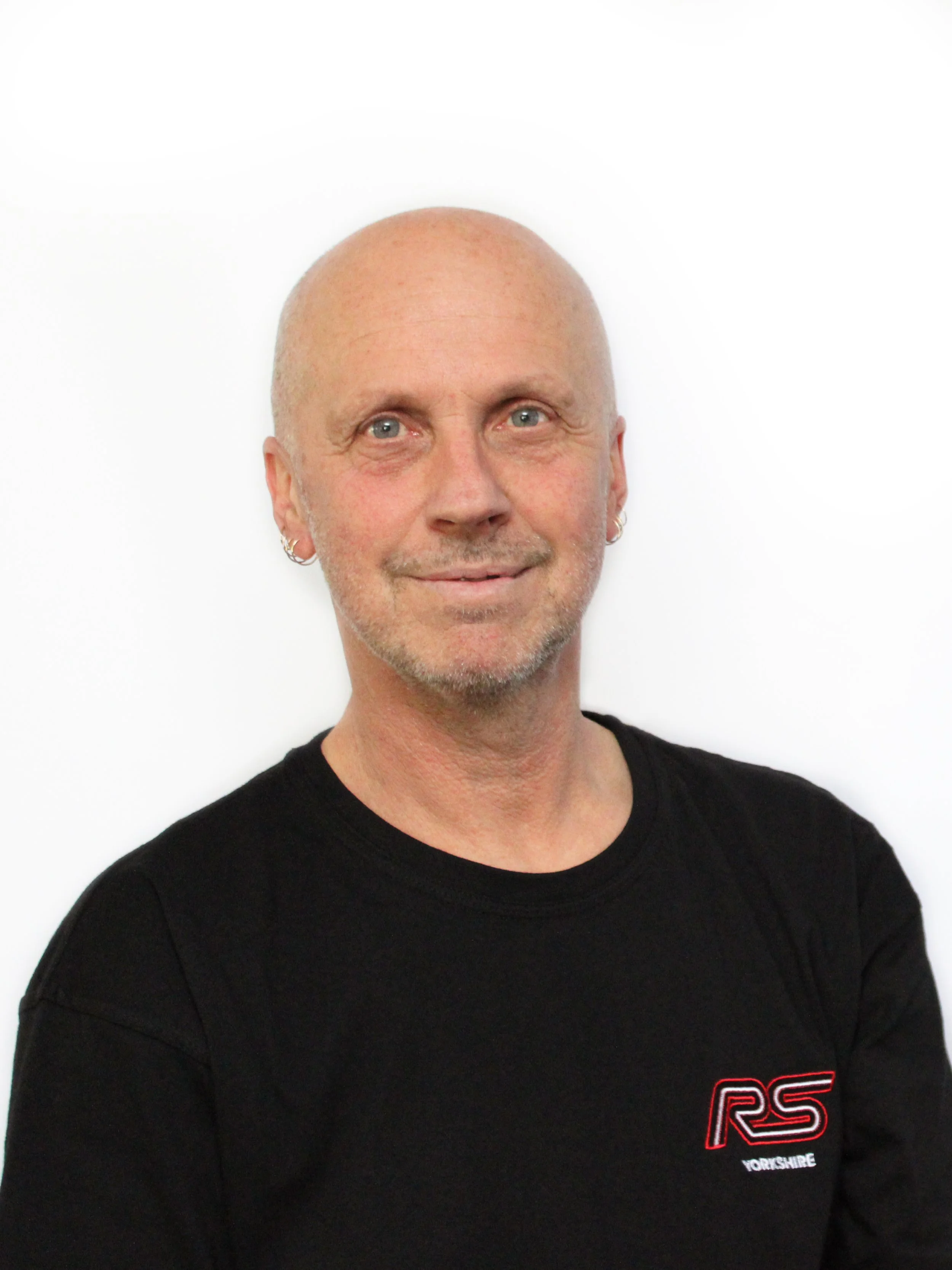 A smiling middle-aged man with a bald head, blue eyes, and earrings, wearing a black shirt with red and white logo, standing against a white background.