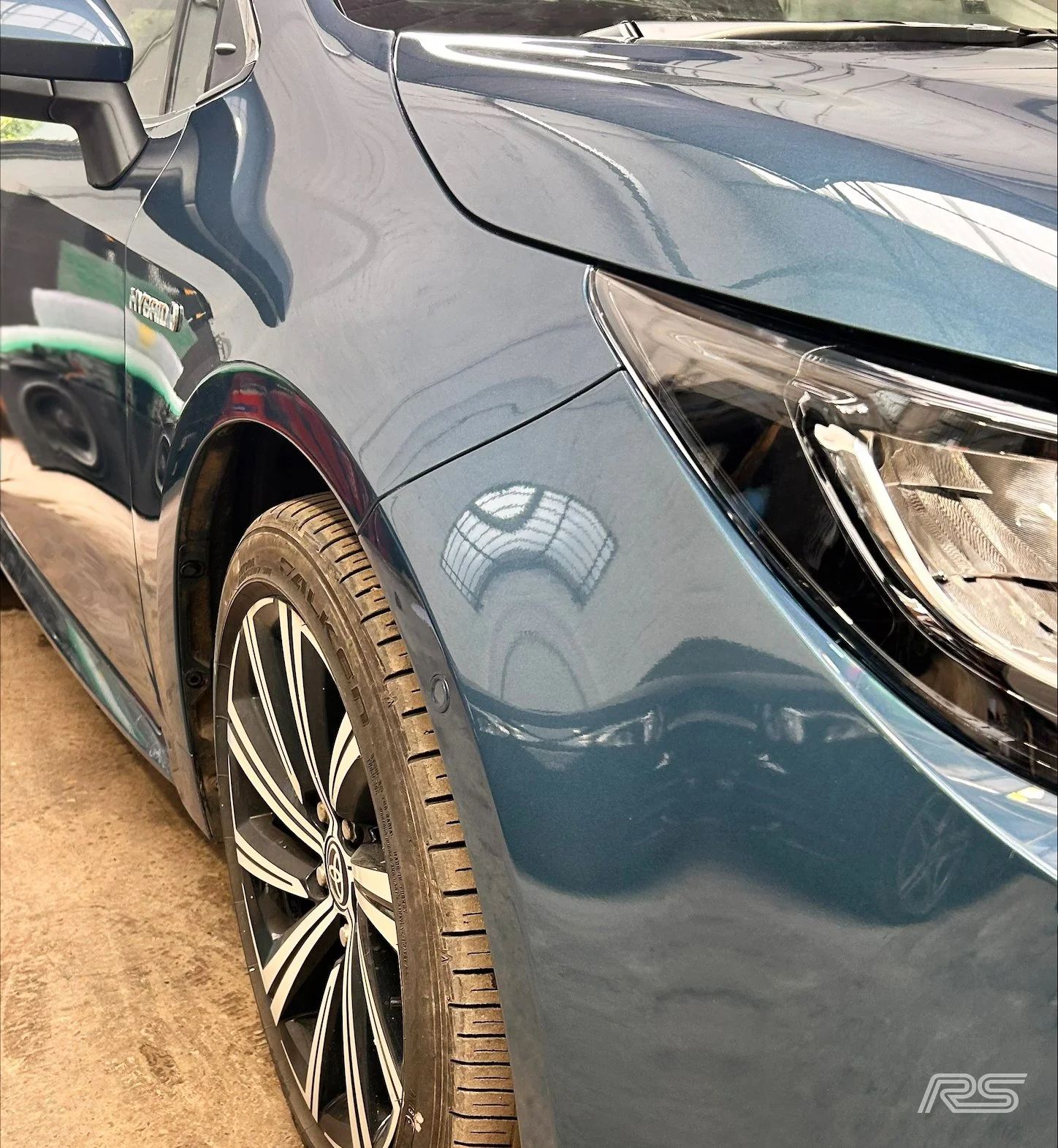 Close-up of the front right side of a blue car, showing the headlight, part of the hood, front wheel, and tire.