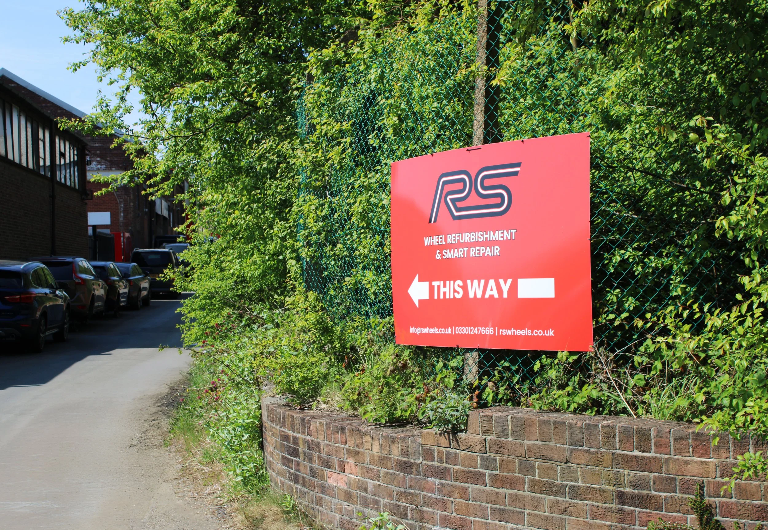 Red sign for RS Wheel Refurbishment & Smart Repair with white arrow pointing left and the text "This Way" in an outdoor setting.
