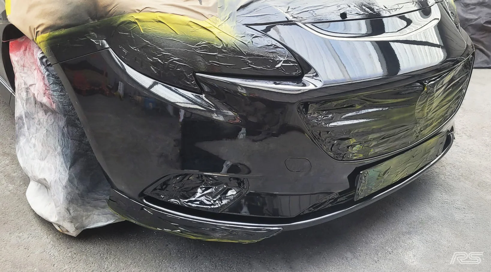 The front of a black sports car covered with a protective plastic wrap, midway through an onsite repair.