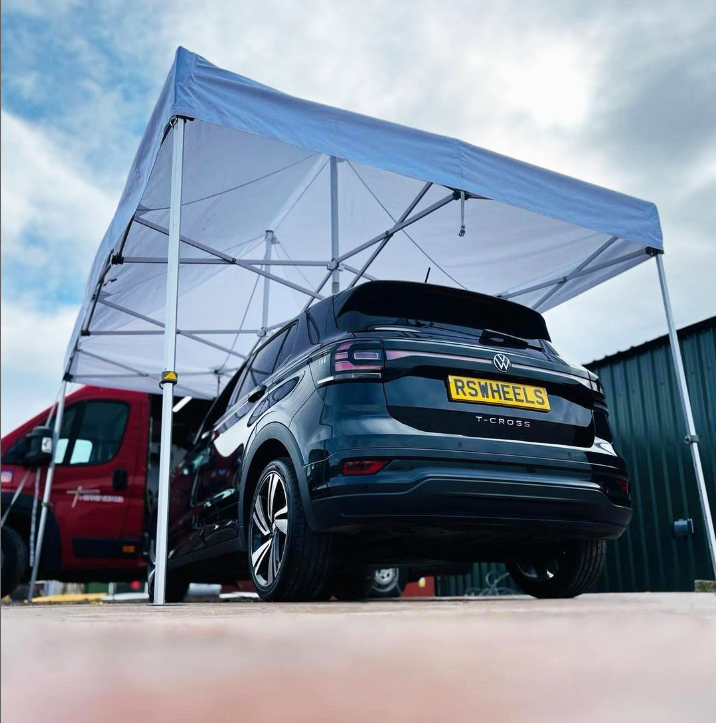 Black Volkswagen T-Cross parked under a white portable canopy with a red vehicle nearby and a green building in the background.