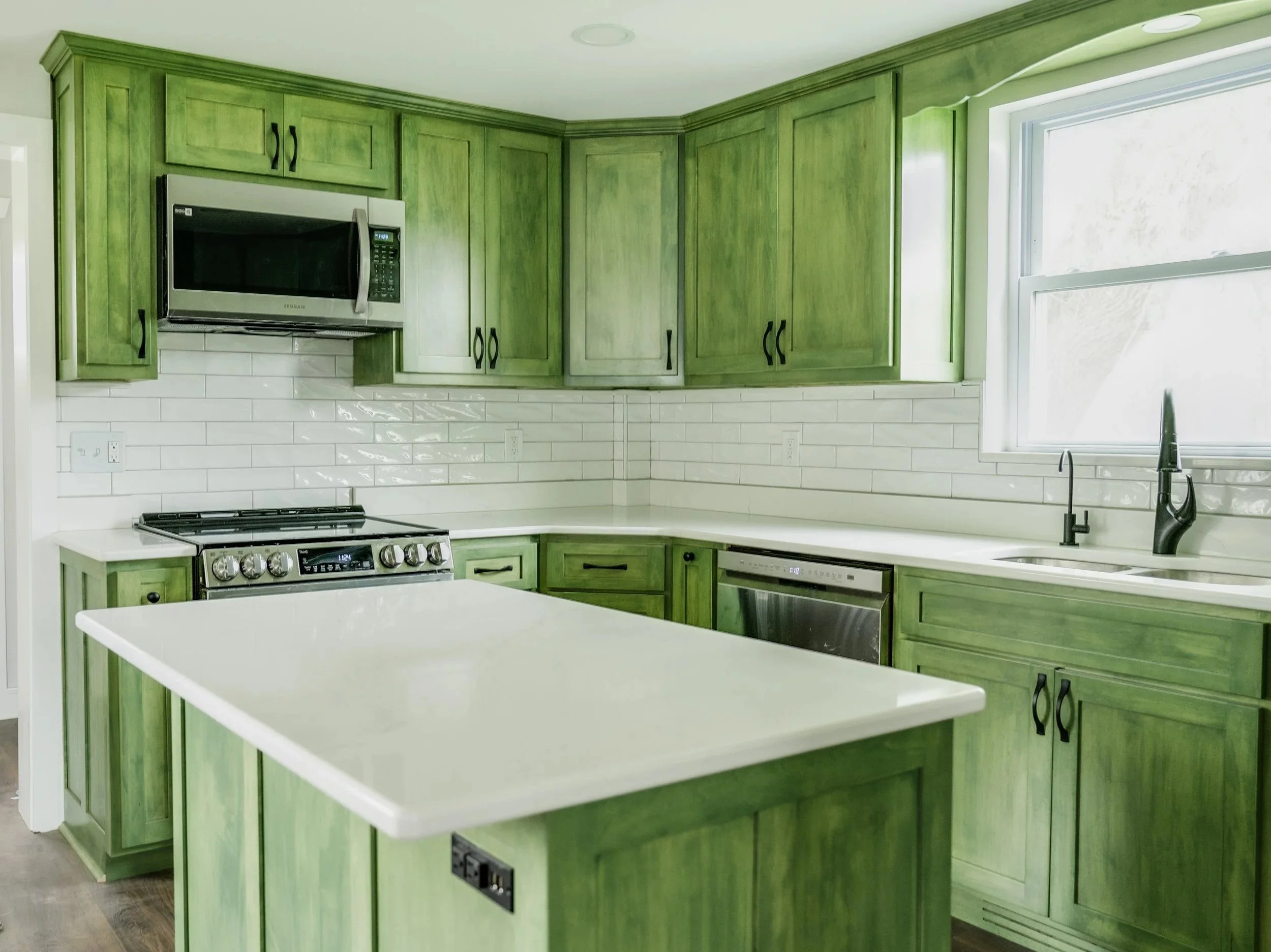 Custom Green-stained kitchen cabinets with white countertops, stainless steel microwave, oven, and dishwasher, white subway tile backsplash, and a window above the sink.