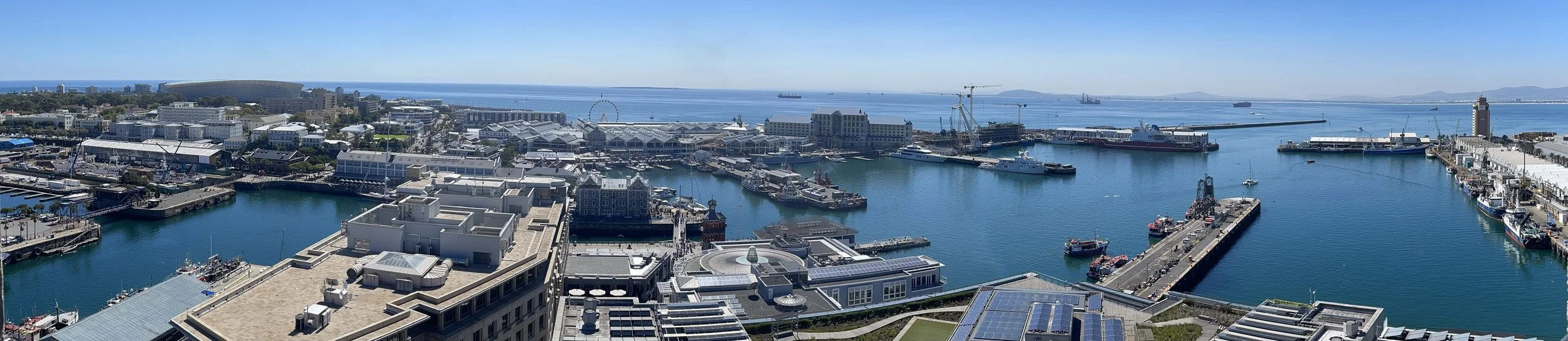 A panoramic view of Cape Town's waterfront area, including the bay, docks, ships, and city skyline under a clear blue sky.