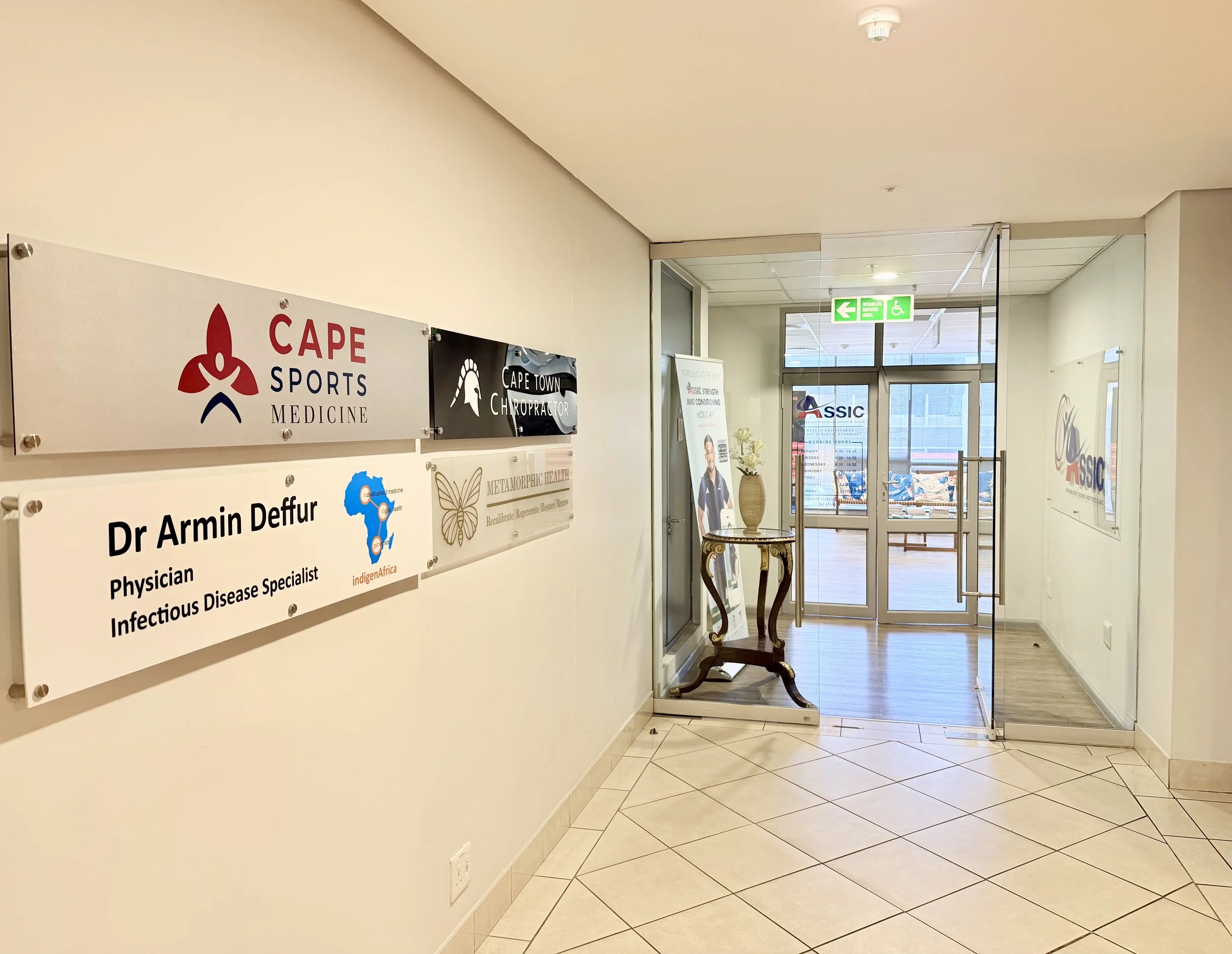 Indoor view of a hallway with several signs, including one for  Dr. Armin Deffur. Glass doors at the end lead to another room with seating and an exit sign.