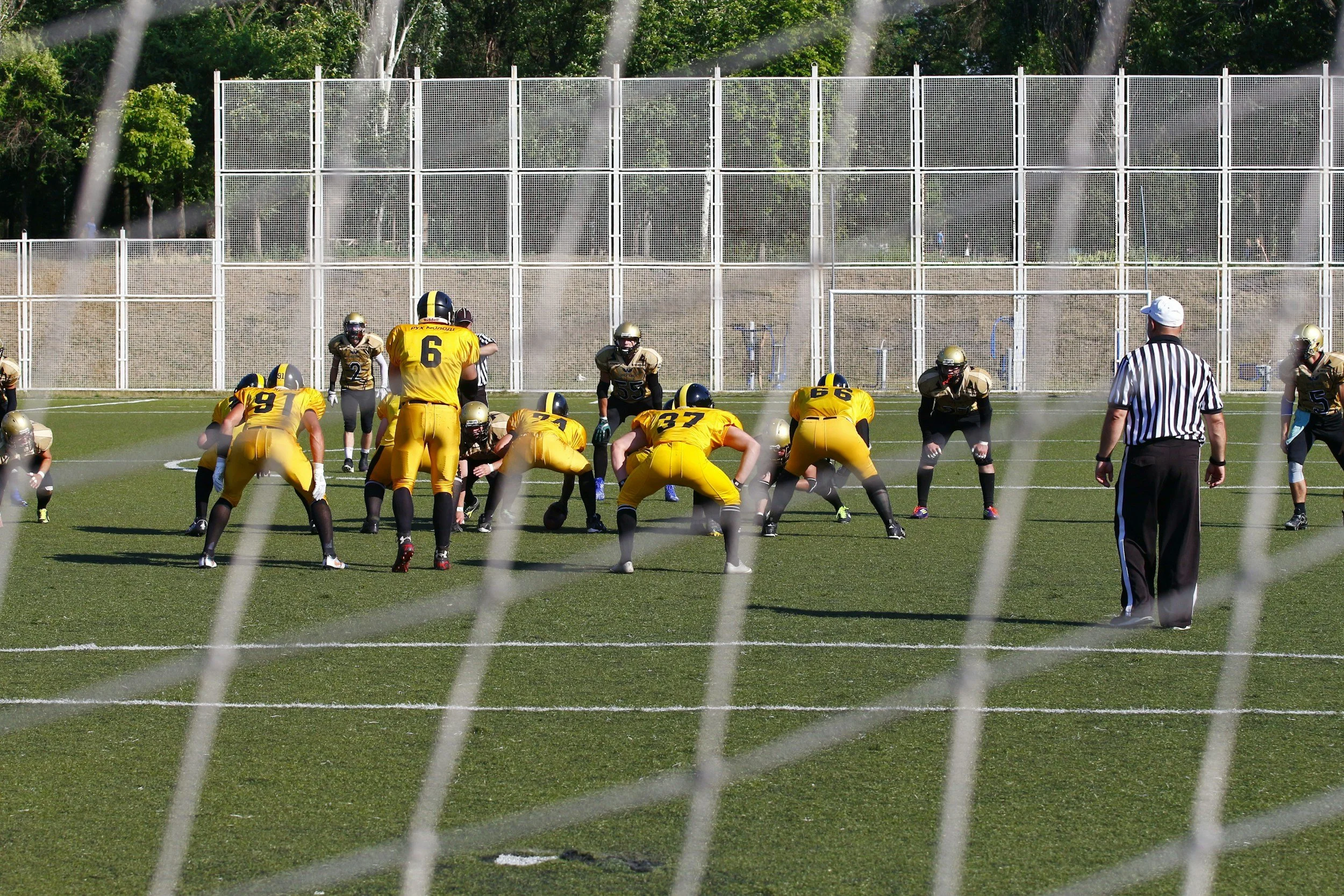 American football game viewed through a goal net, with players in yellow and black uniforms facing each other on the field, and an official standing nearby.