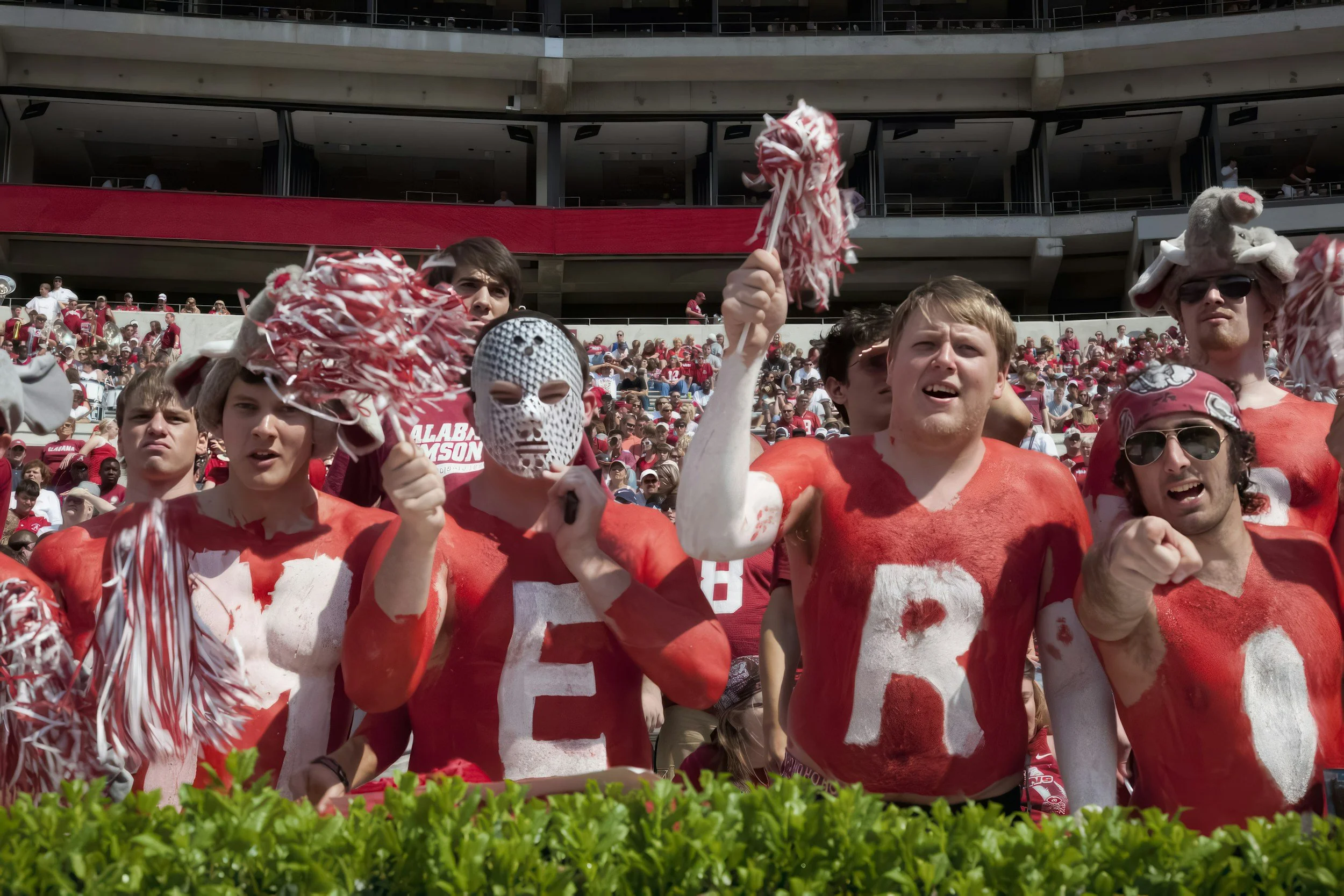 Crowd of football fans dressed in red shirts with painted letters, cheering at a stadium. Some fans are wearing masks and hats, holding pom-poms.