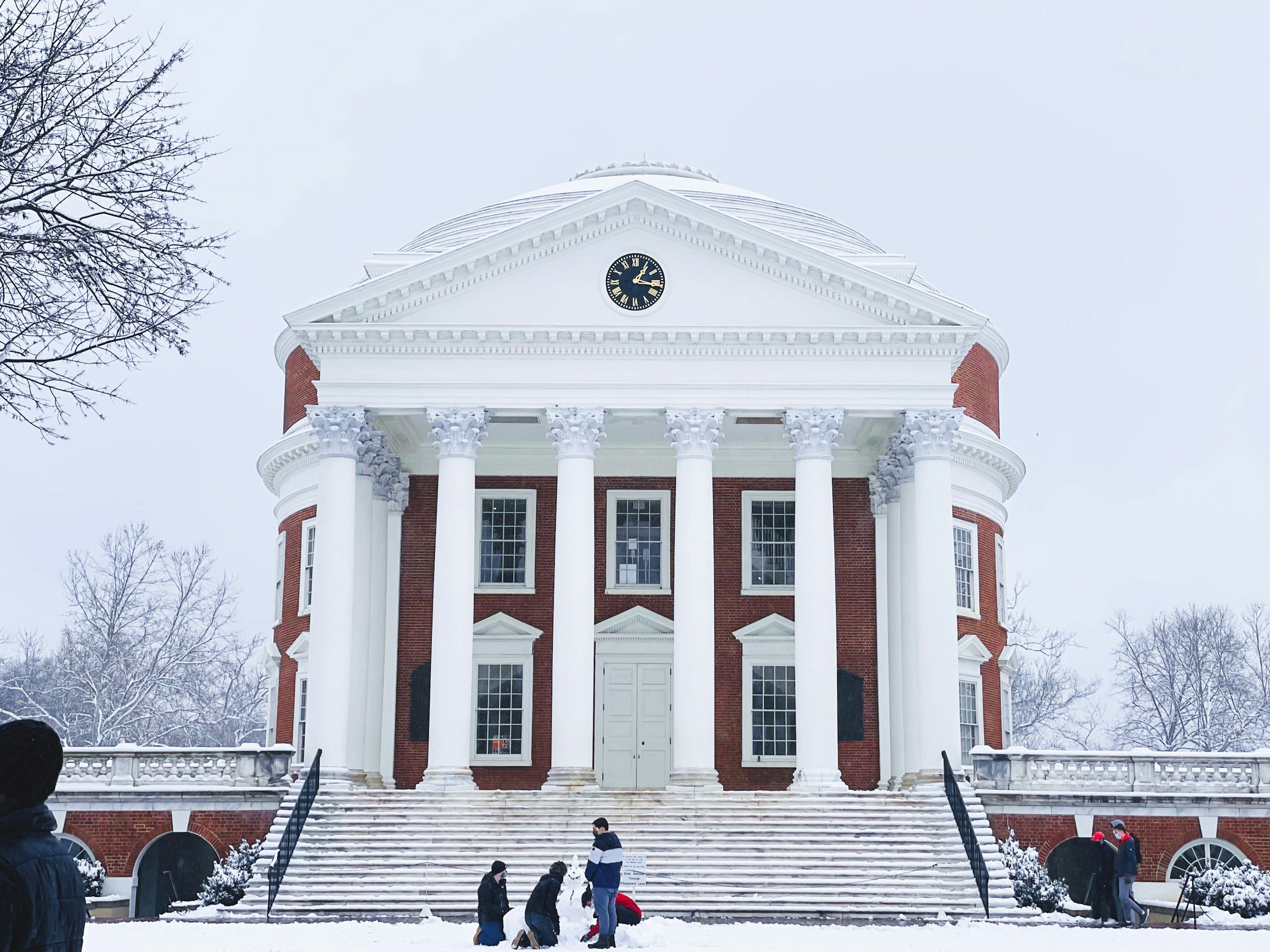 A large historic building with a white classical façade, tall columns, and red brick walls, covered in snow during winter, with a group of people in front.