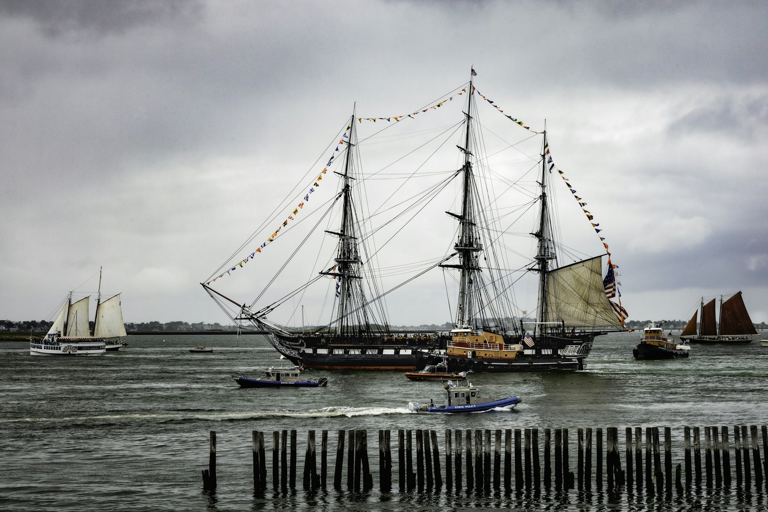 Large black sailing ship decorated with signal flags, surrounded by smaller boats and yachts on the water, with a pier or wooden posts in the foreground and a cloudy sky in the background.