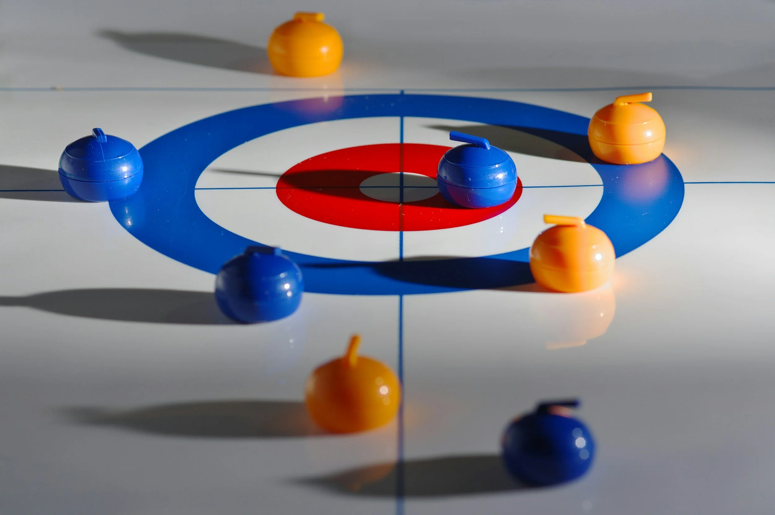A curling game in progress with blue and yellow stones on a white curling sheet with red and blue rings and a guiding line.