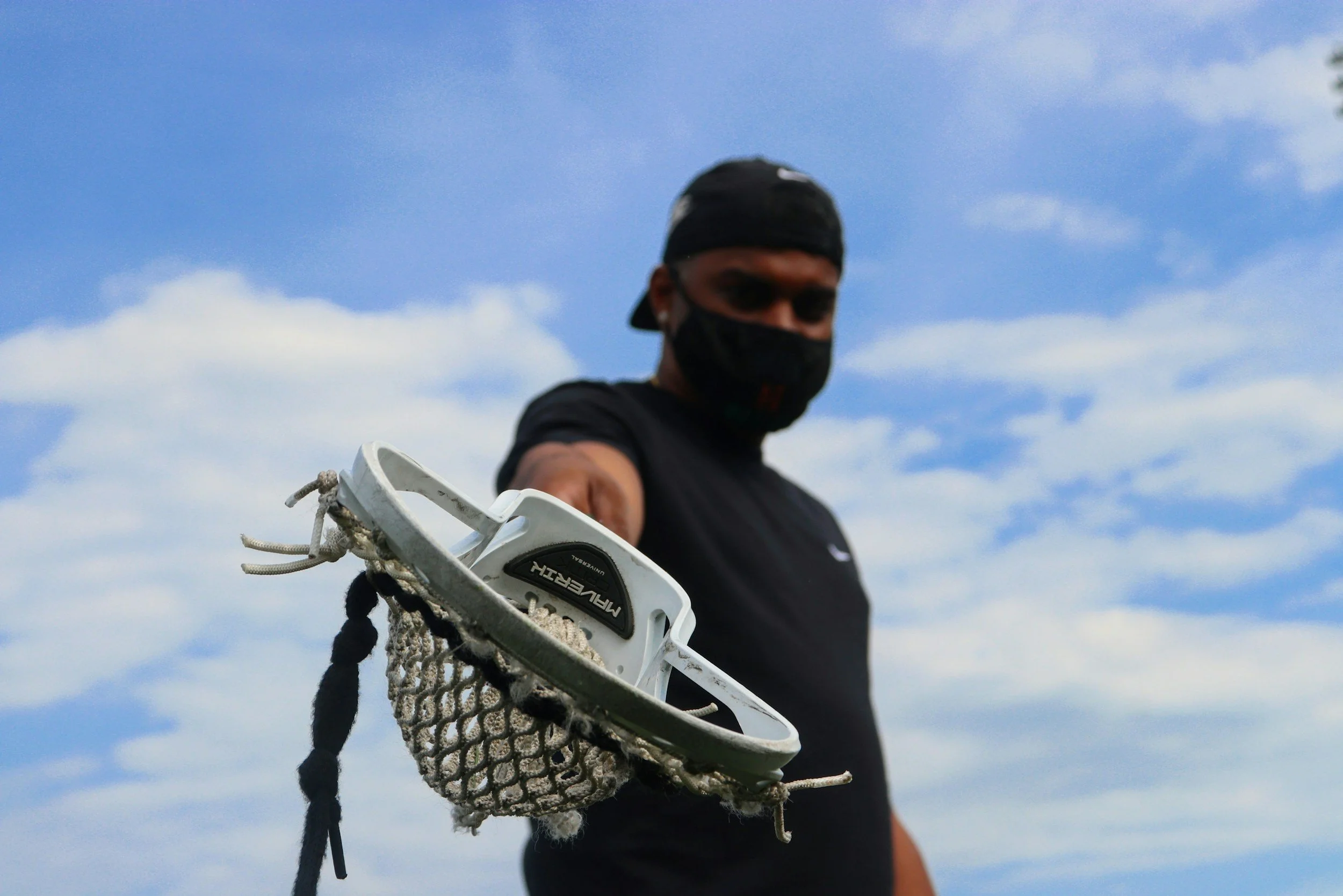Person wearing black shirt and face mask holding a lacrosse stick towards the camera outdoors
