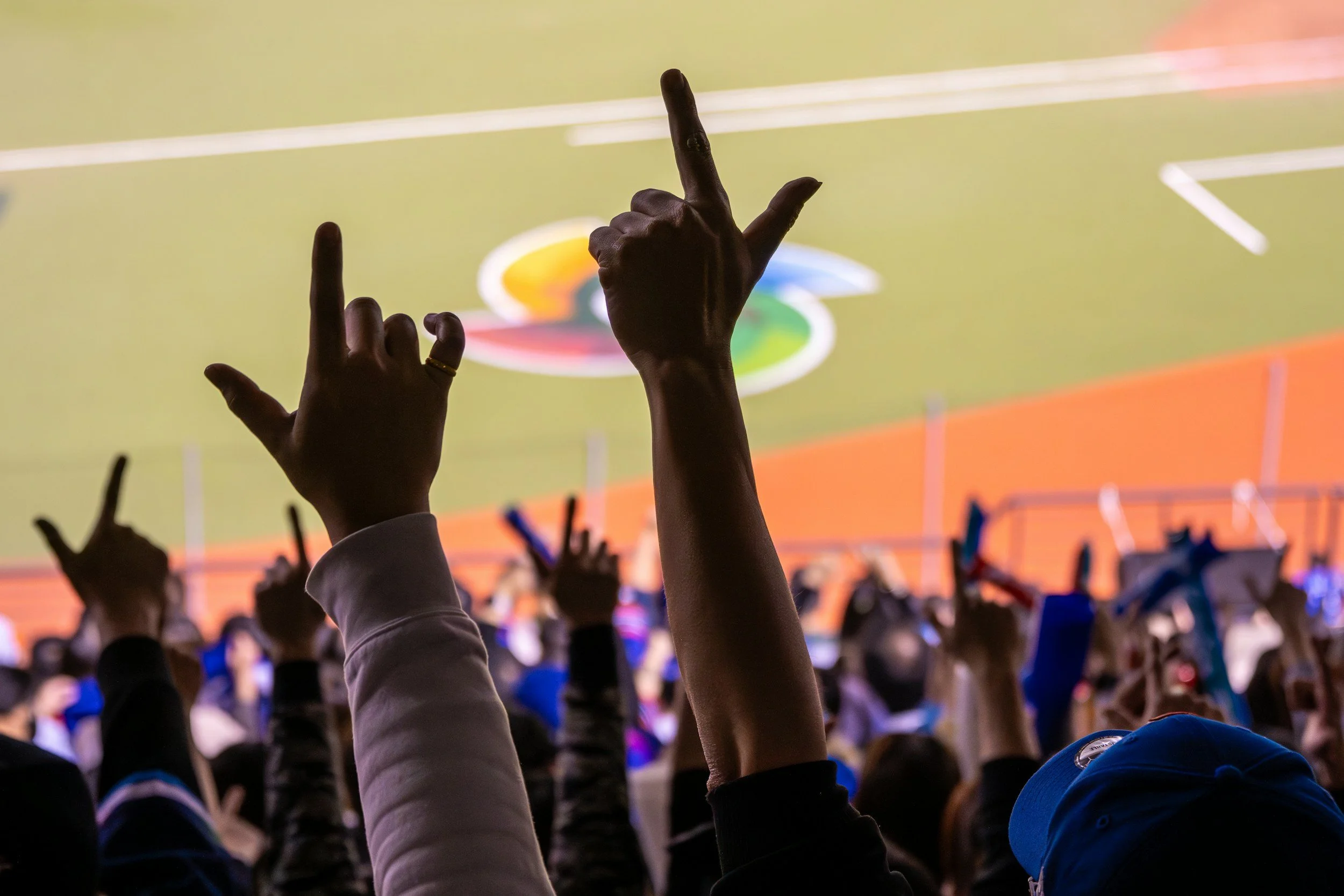 Crowd at a sports event raising their hands with some forming the sign of the horns, with a brightly lit basketball court in the background.