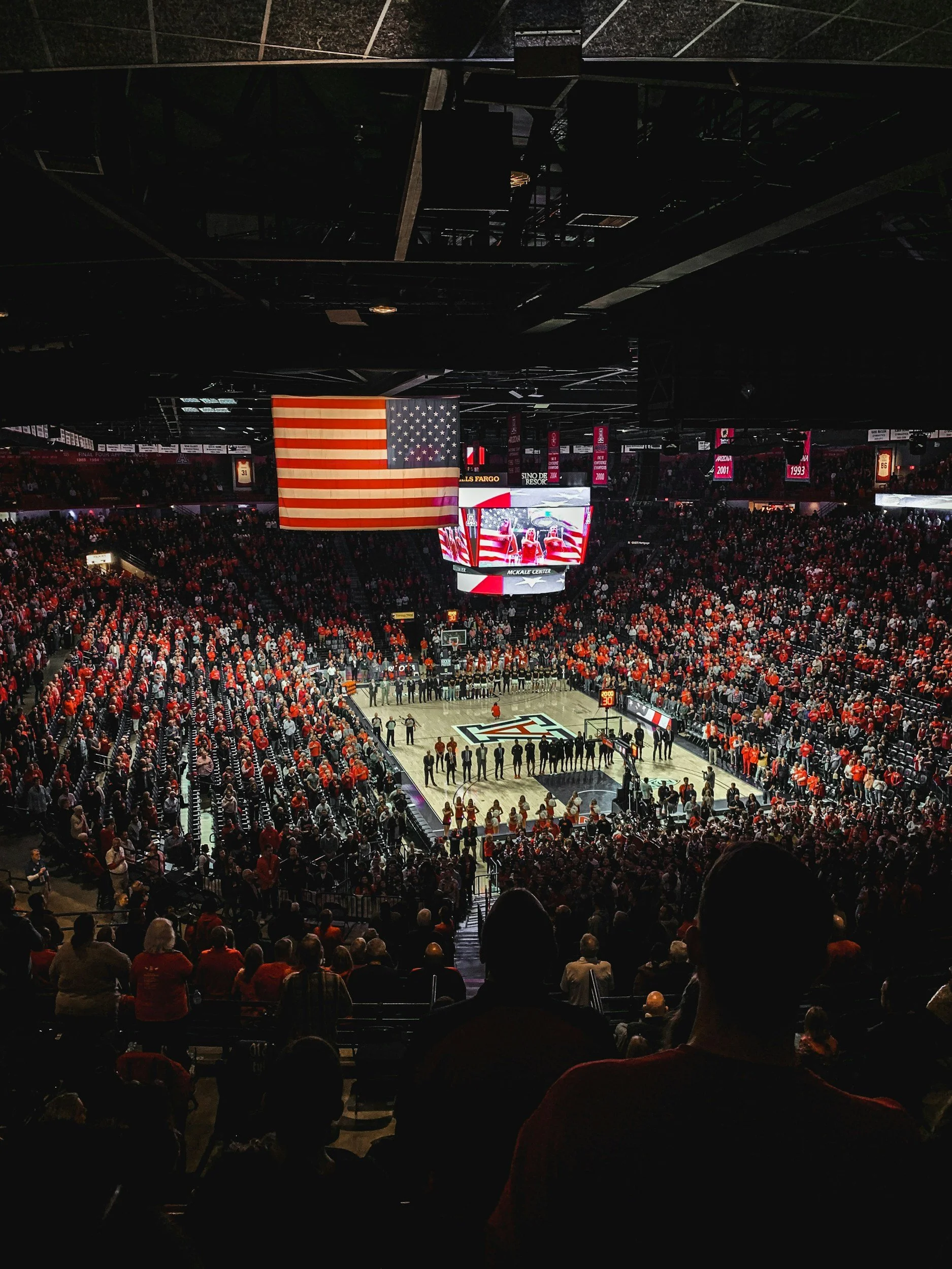 Crowded basketball arena with American flag hanging above the court, surrounded by fans and banners, showing an event or game.