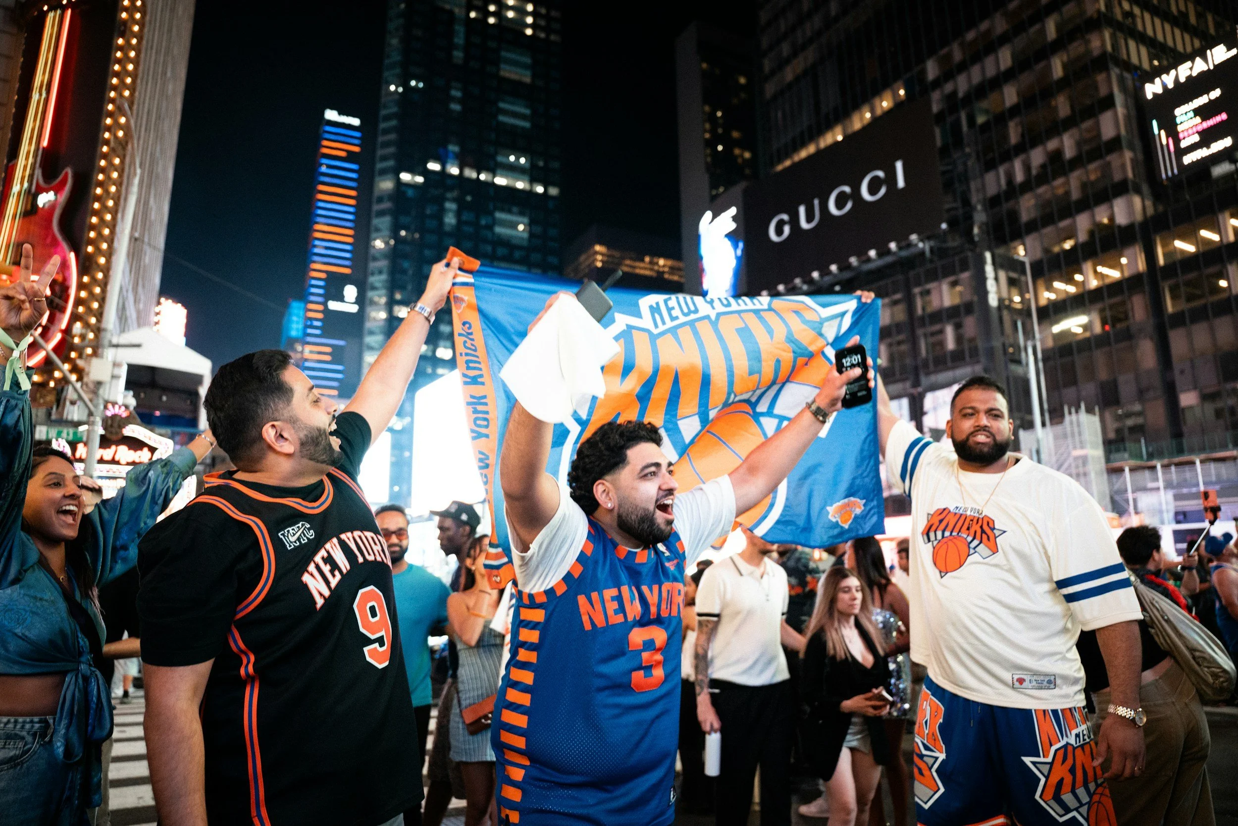 Group of people celebrating New York Knicks basketball game in Times Square at night, holding a Knicks banner, wearing team jerseys, surrounded by bright signs and tall buildings.