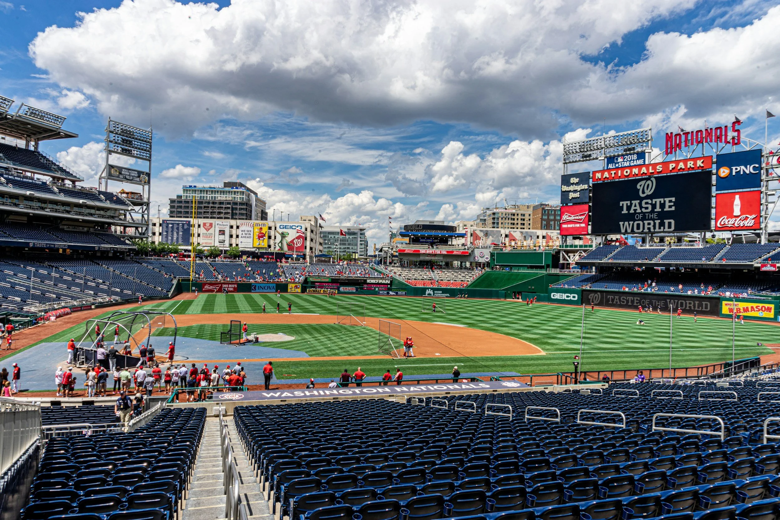 Empty baseball stadium with players warming up on the field, bright sky with clouds, and large electronic scoreboard displaying 'Taste of the World' at Nationals Park in Washington, D.C.