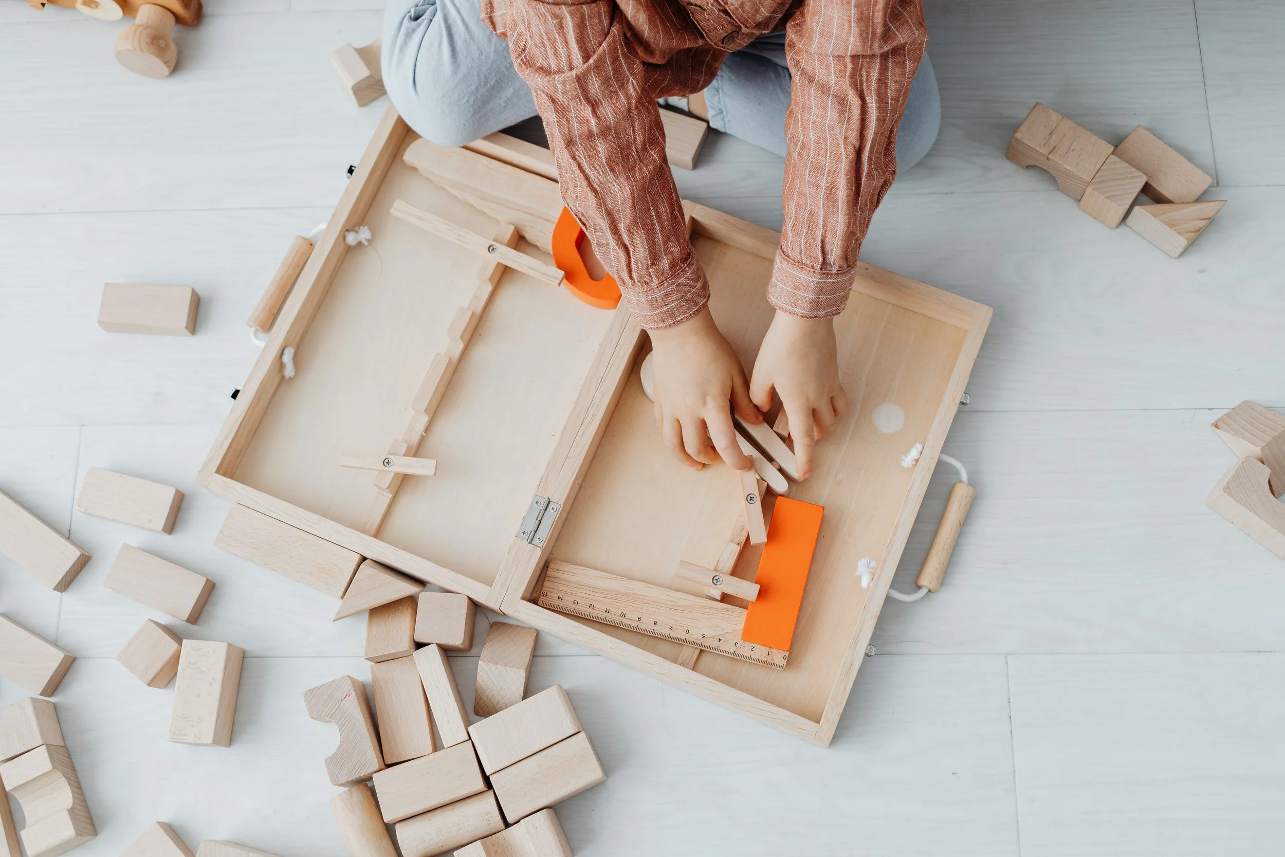 A person assembling a wooden block structure on a light-colored floor, surrounded by scattered wooden blocks.