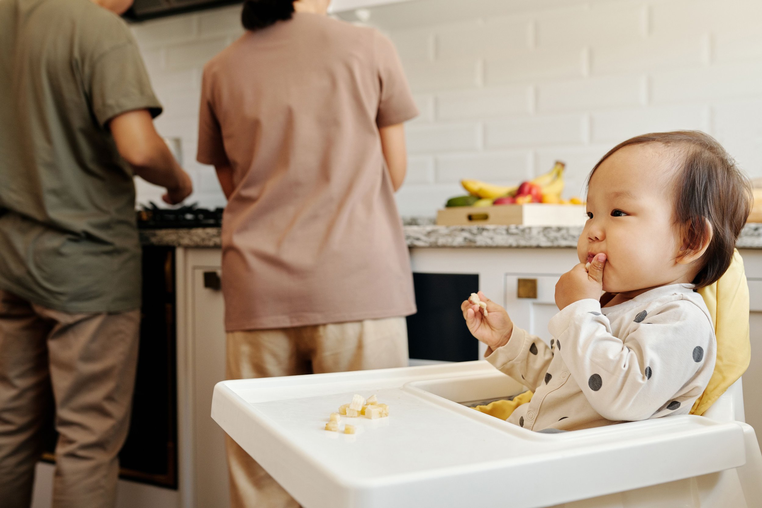 A young child sitting in a high chair, eating a snack, with a kitchen in the background. Two adults are standing nearby, one near the stove and the other with their back to the camera. A fruit bowl with bananas and other fruits is on the countertop.