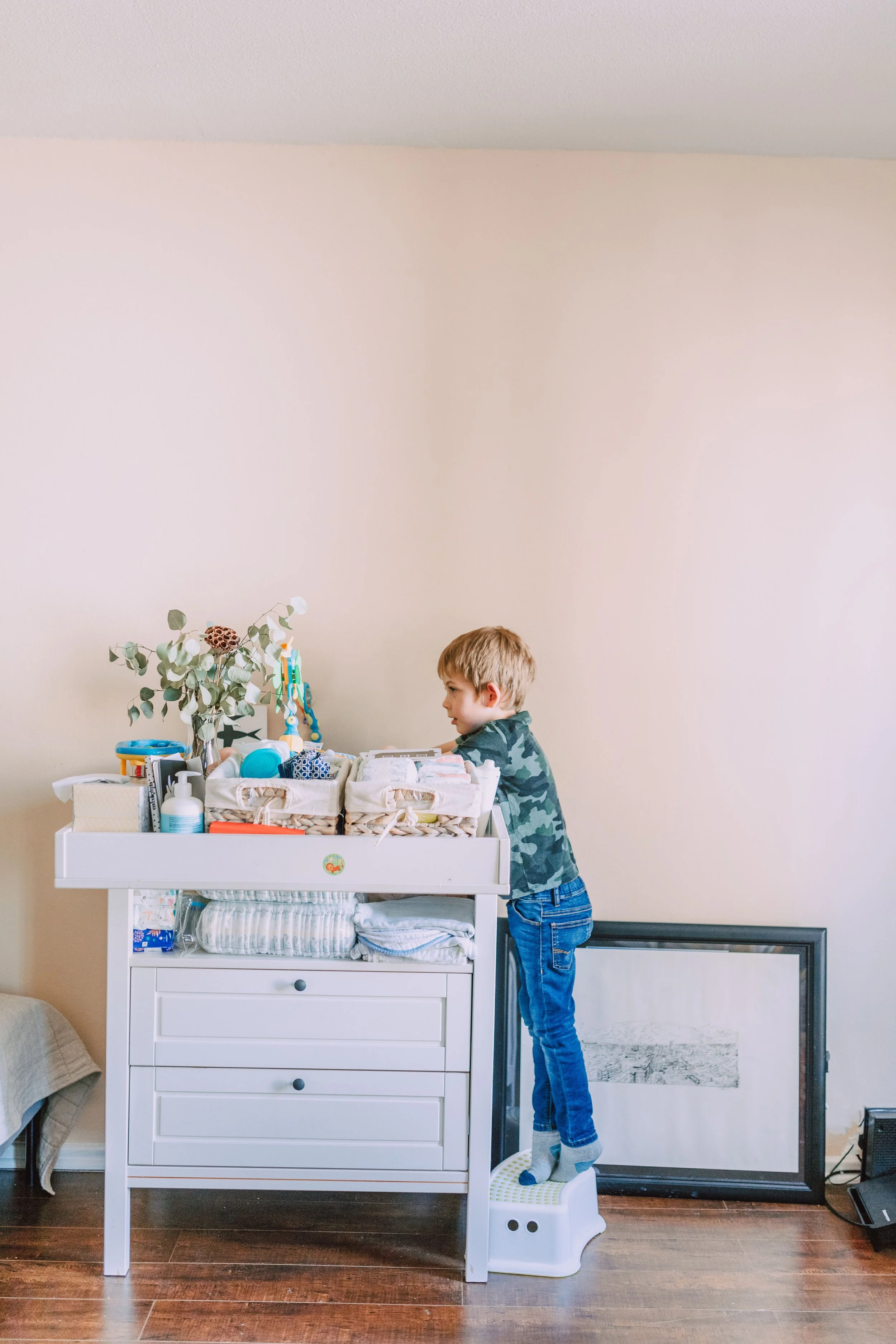 A young boy with blonde hair, wearing a camouflage shirt and blue jeans, stands on a small step stool in front of a white changing table. The changing table is filled with various items, including a plant with eucalyptus leaves, bottles, and boxes. Behind the boy, there is a beige wall and a large picture frame on the floor leaning against the wall. To the right, a black flat-screen TV is also visible.