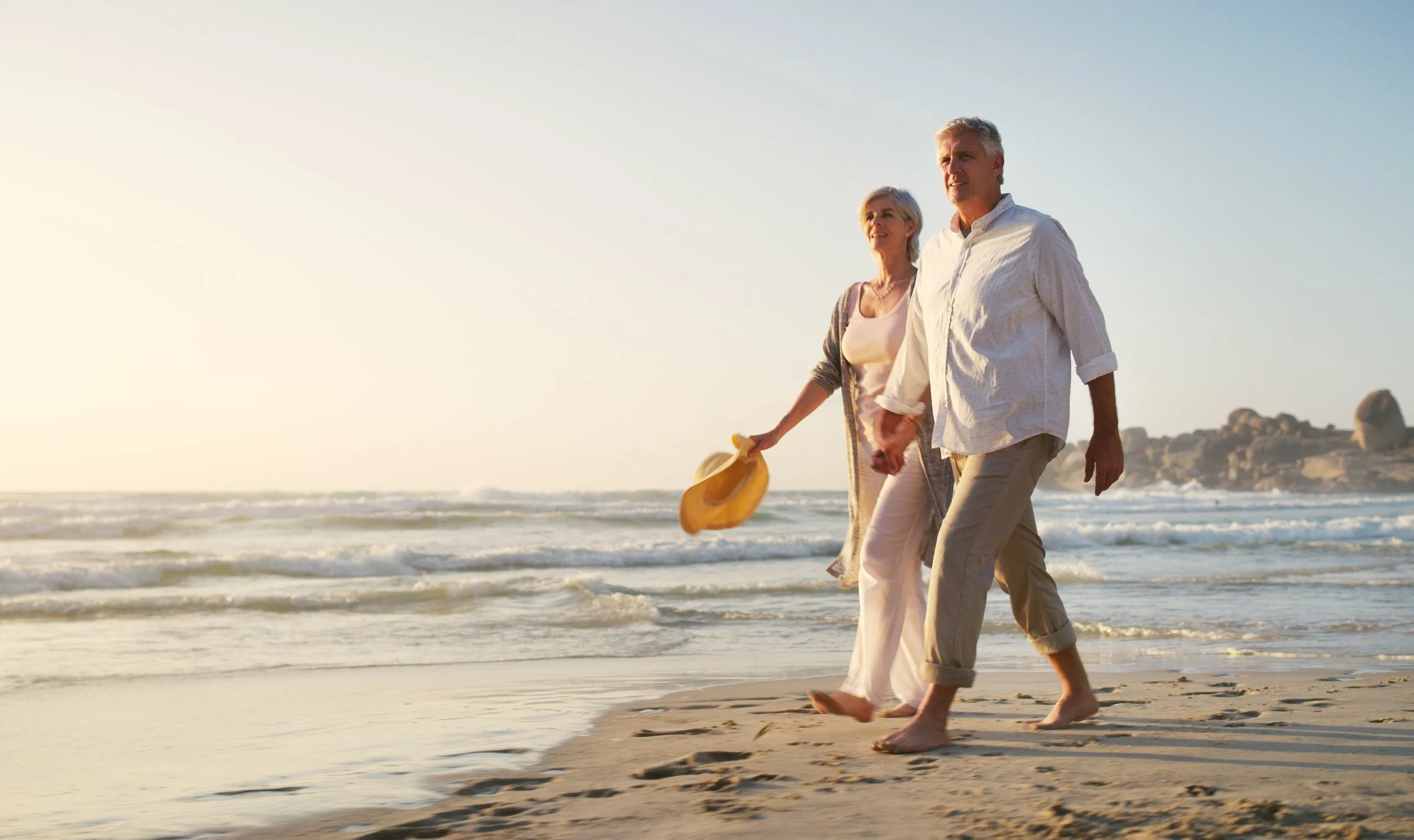 An elderly couple walking hand-in-hand along the beach during sunset, dressed in light clothing, with the woman holding a yellow hat, enjoying a peaceful moment by the ocean.
