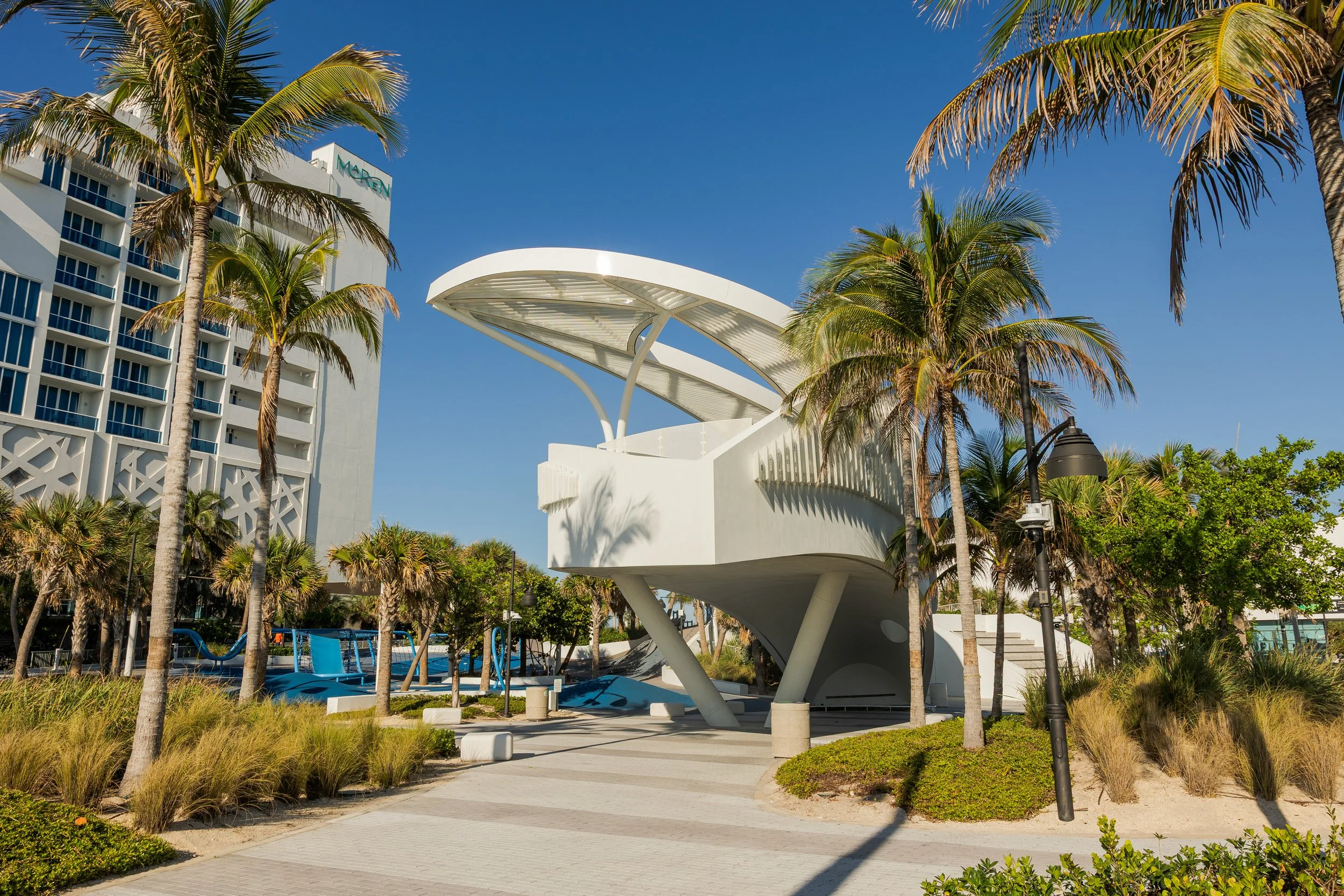 Modern building with a curved white architectural canopy, surrounded by palm trees and a park area with walking paths, lamp posts, and a playground in the background.