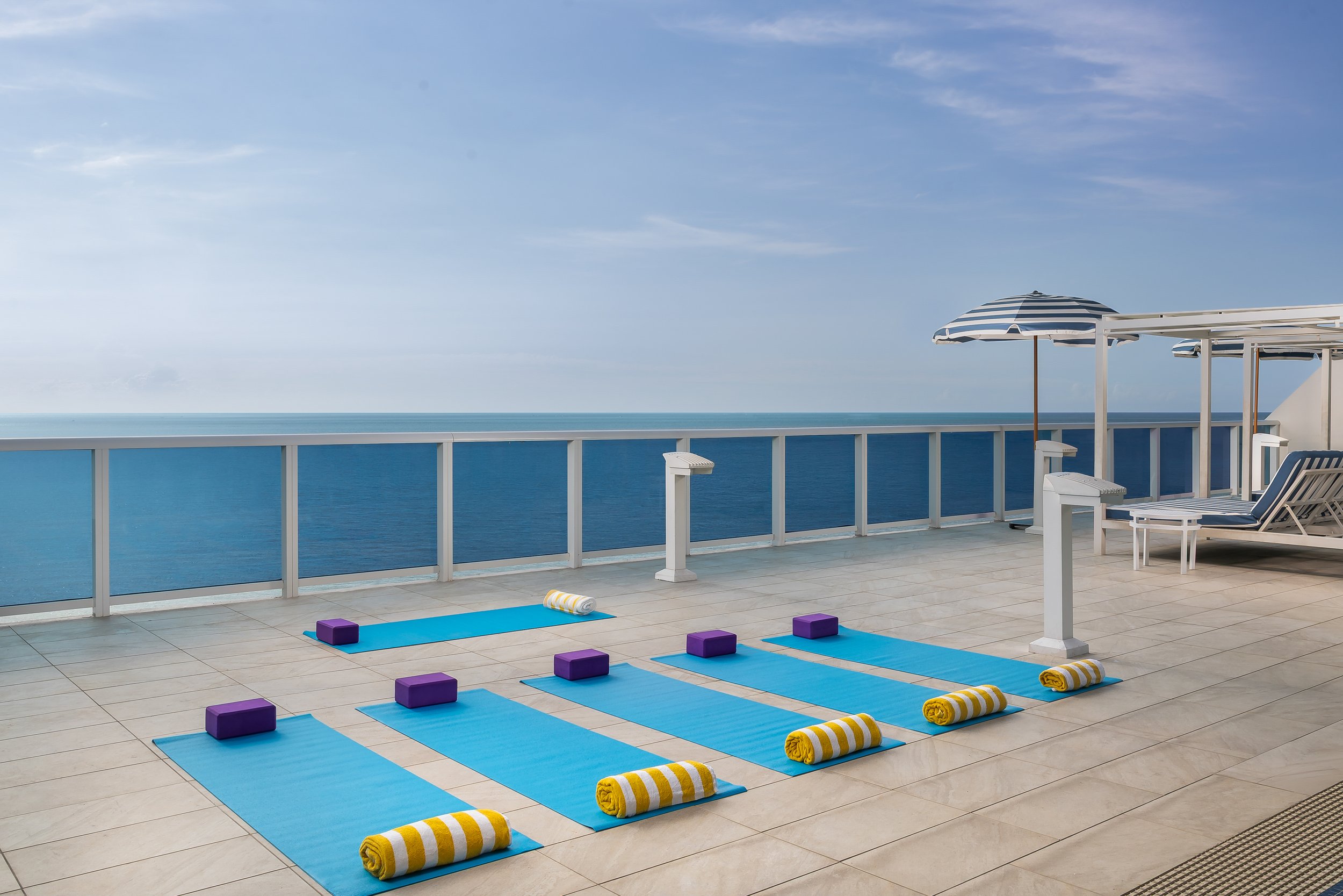 An outdoor balcony with a light-colored tile floor, blue yoga mats with rolled yellow and white striped towels, and purple yoga blocks, overlooking the ocean under a cloudy sky.