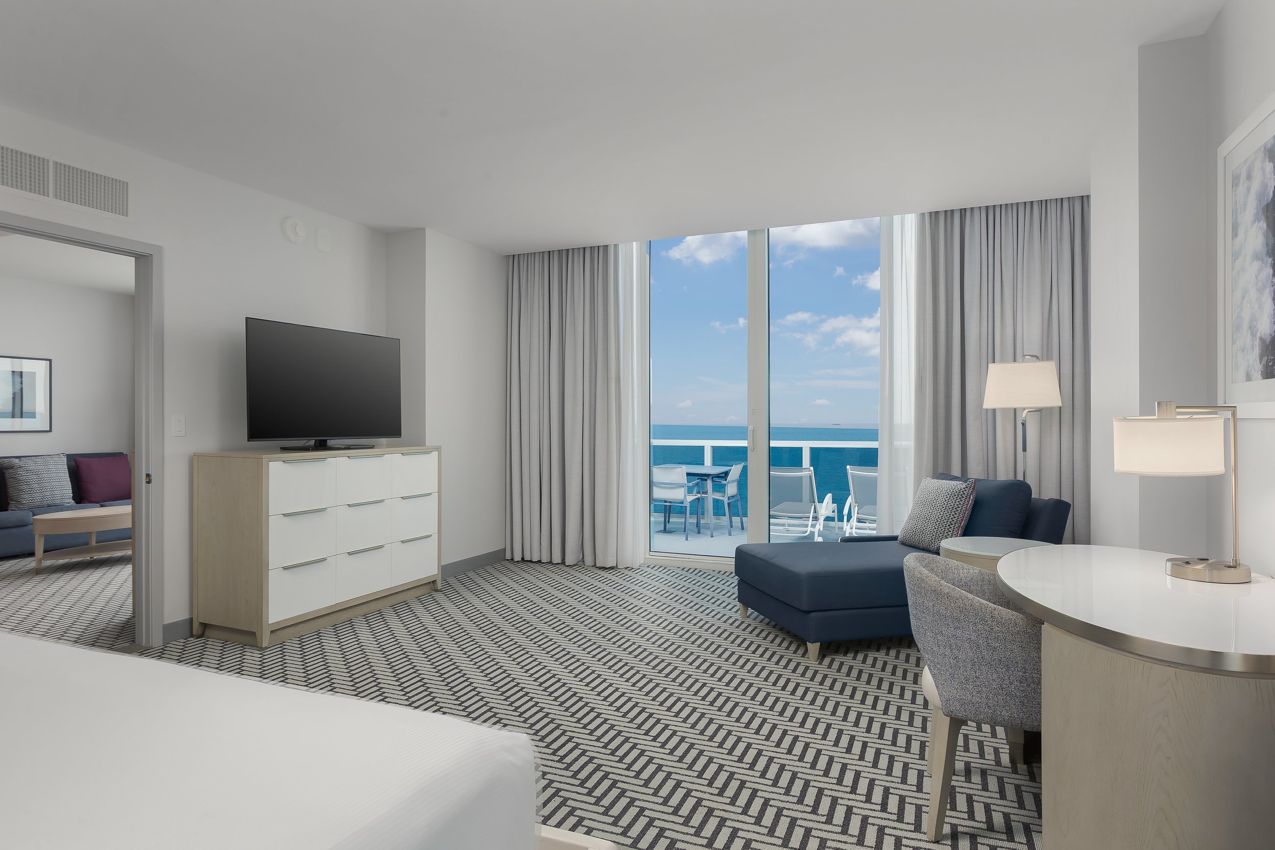 Interior of a modern seaside apartment with white walls and large windows looking out onto an ocean view. The room features a dark blue sofa, a white dresser, a mounted flat-screen TV, lamps, and a round dining table. Curtains frame the windows, and 