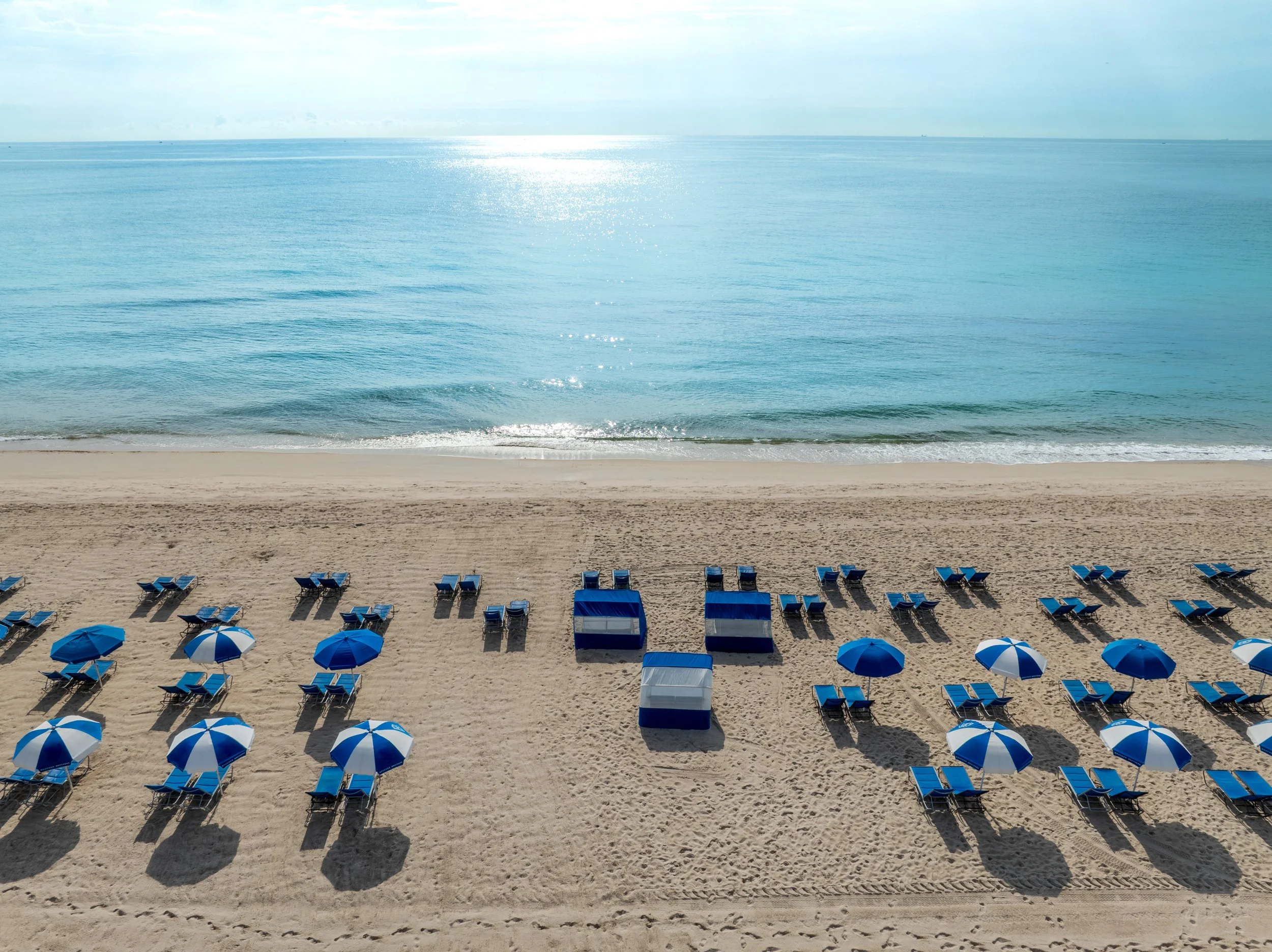 Empty beach with blue and white striped umbrellas and chairs, calm ocean waves, and a bright sky with some clouds.