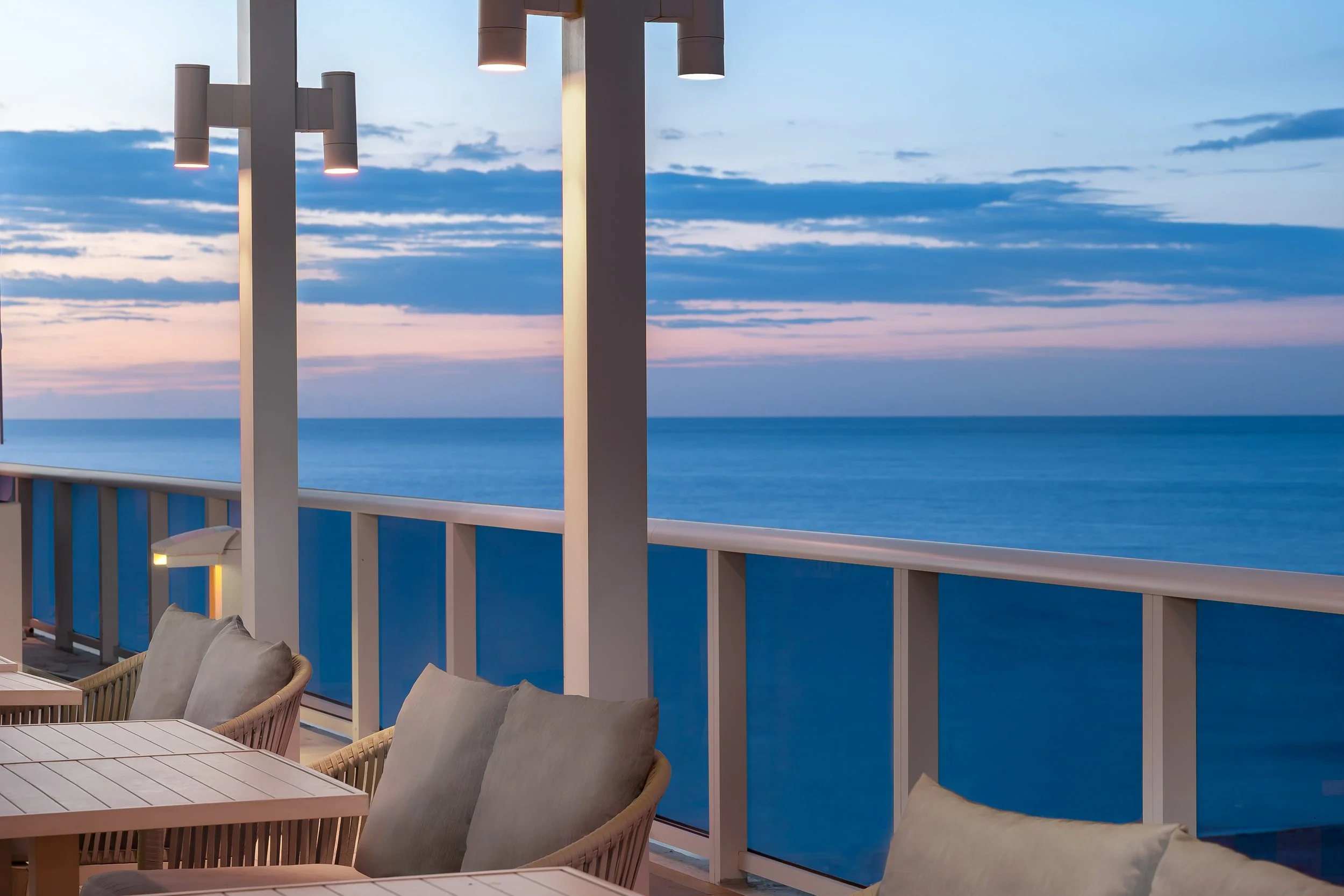 Balcony with outdoor seating overlooking the ocean at sunset, with wooden tables, cushioned chairs, and a railing, in a coastal resort setting.