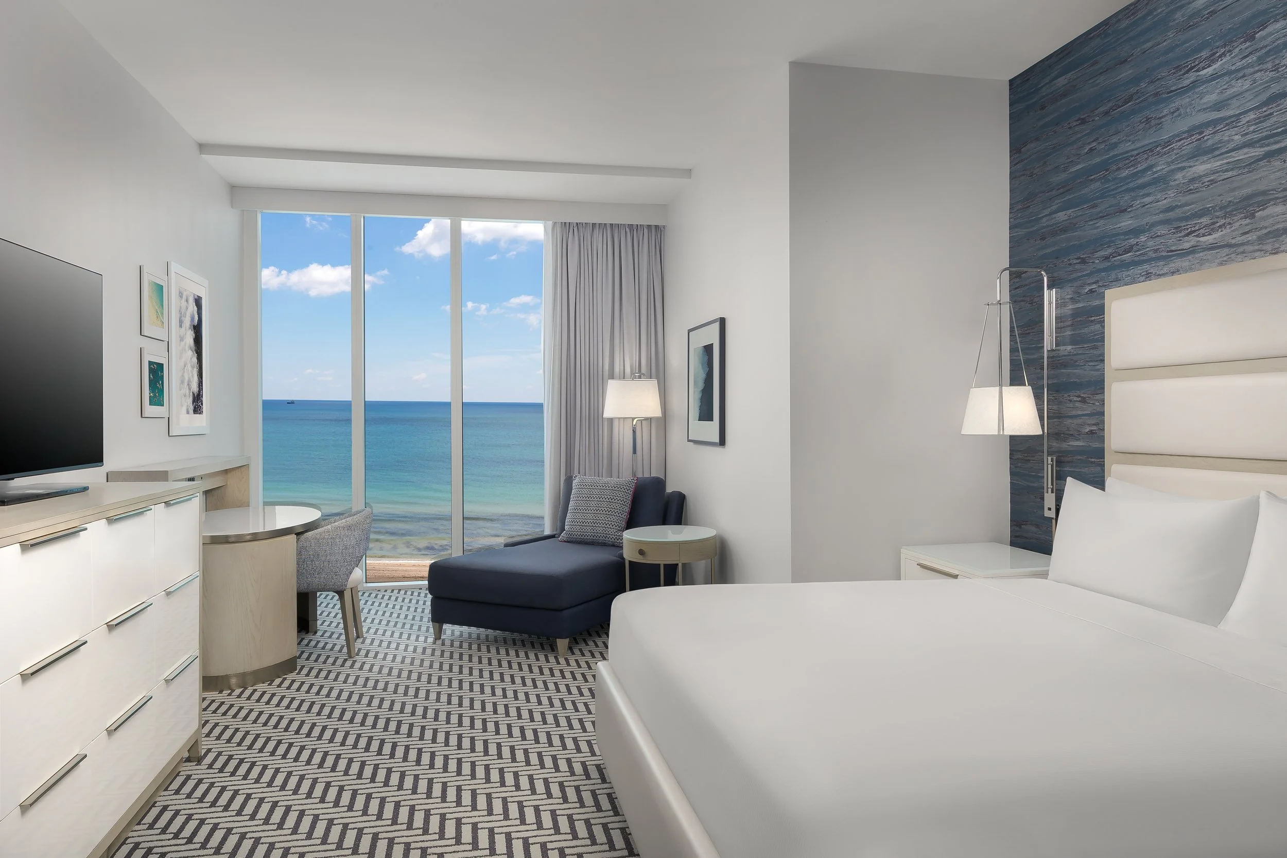 Modern hotel room with white walls, large windows showing the ocean and blue sky, a bed, a TV, and artwork on the walls.