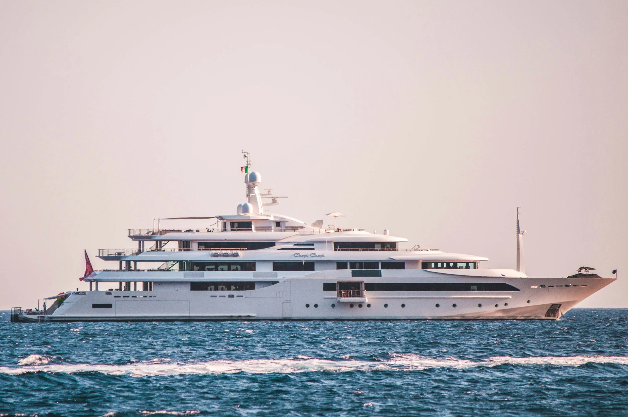 A large white yacht with multiple decks is sailing on the ocean with gentle waves, under a pale sky.