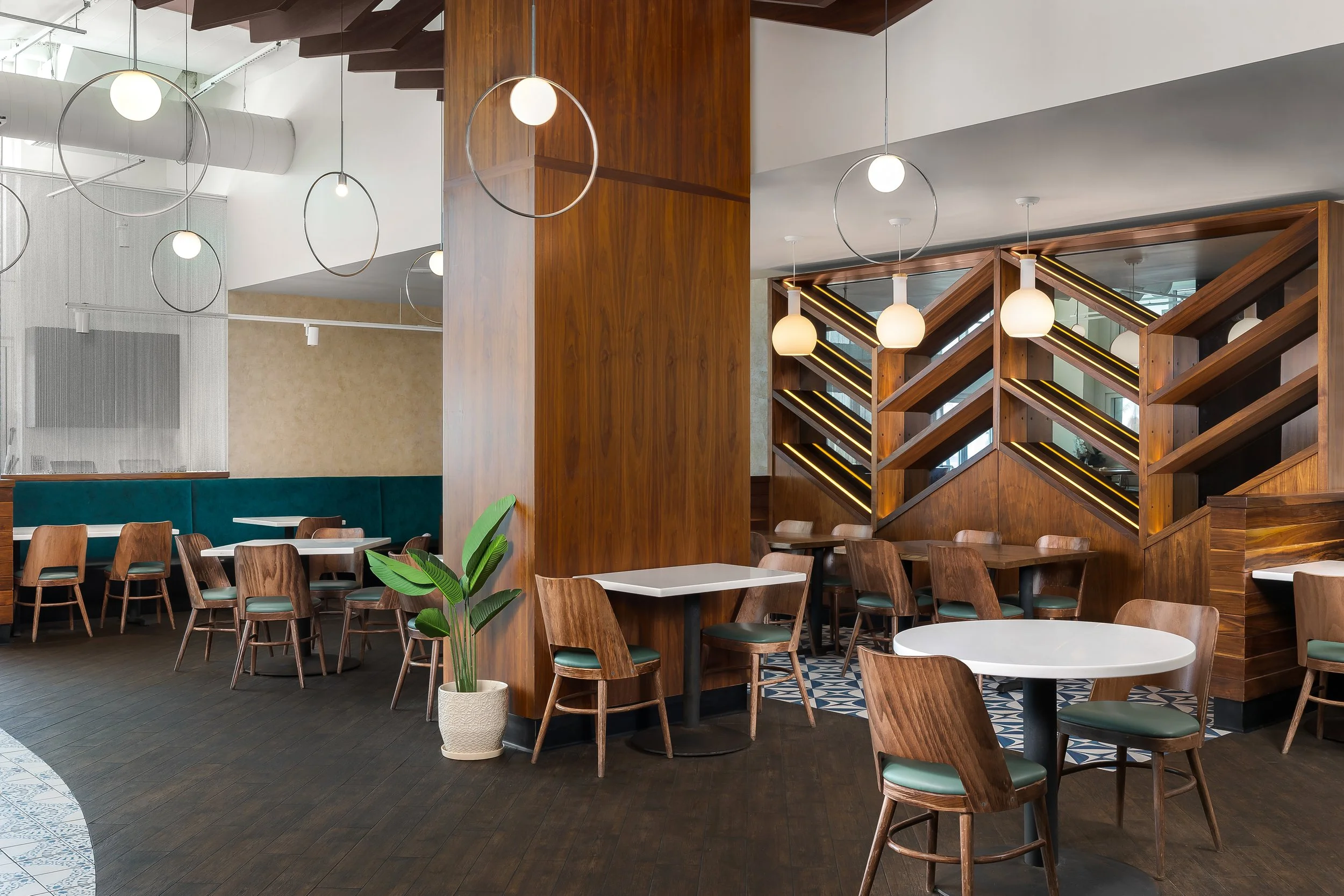 Modern restaurant interior with wooden accents, hanging pendant lights, and a staircase with yellow lighting details. There are tables, chairs, and a large plant in a white pot.