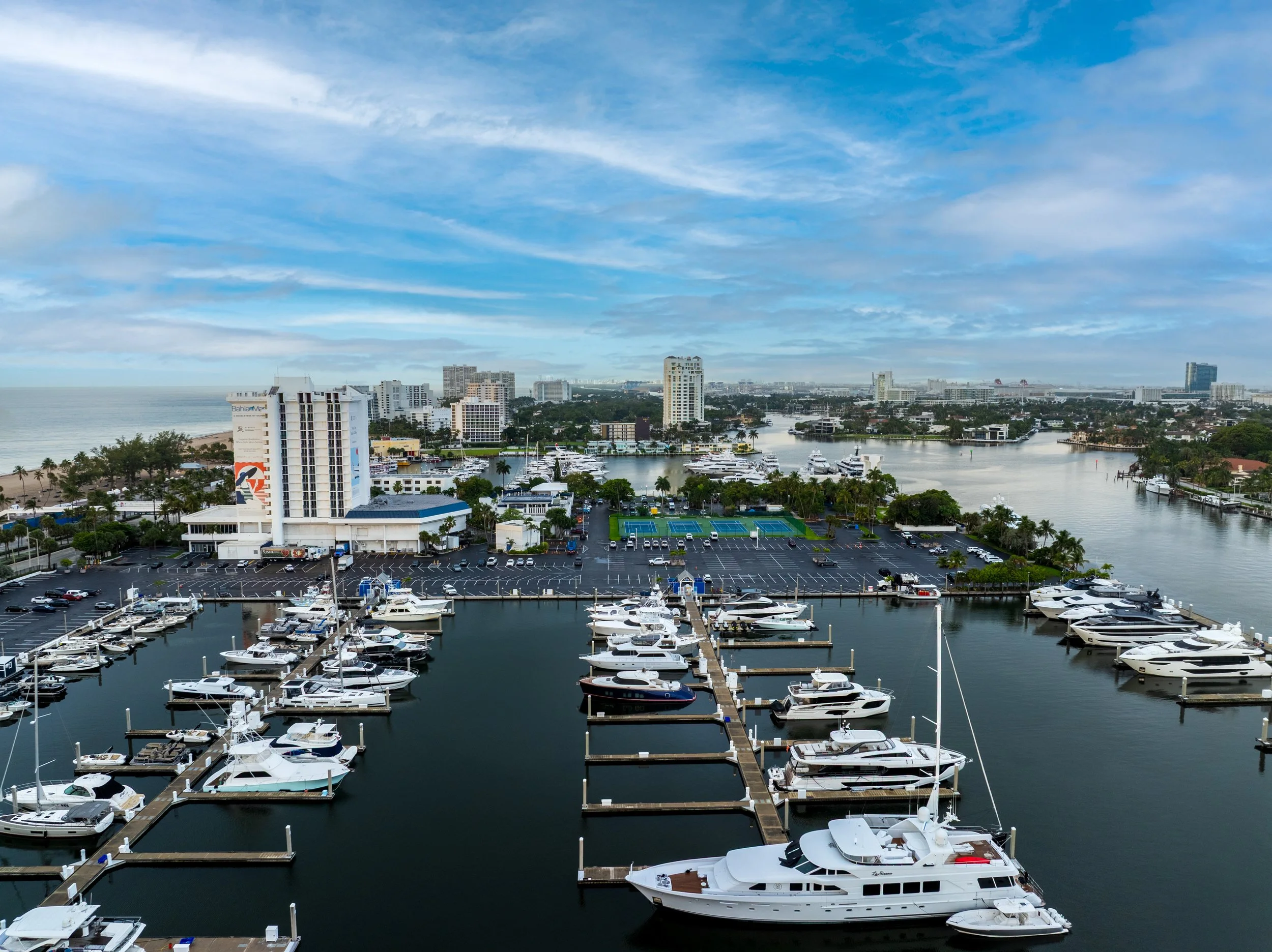 Aerial view of a marina with yachts docked, high-rise buildings, and a city skyline under a partly cloudy sky.
