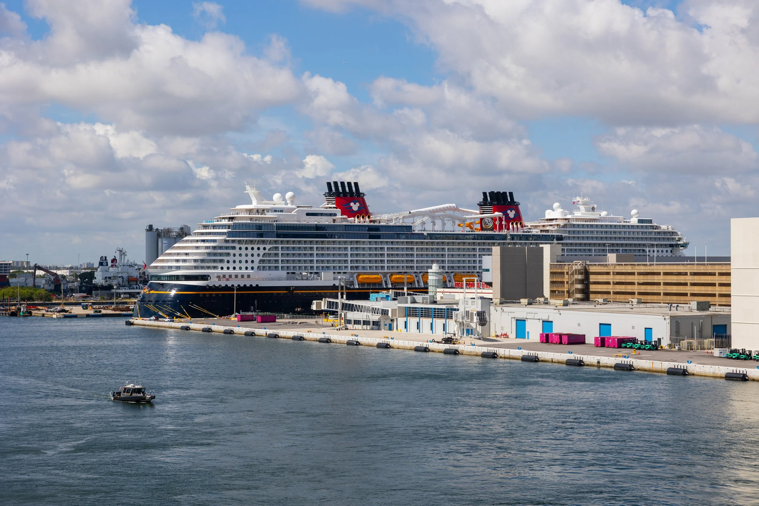 A large Disney cruise ship docked at a port with a harbor in the foreground and cloudy sky above.