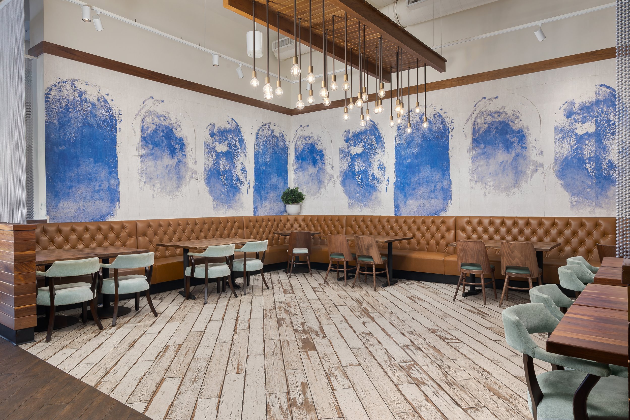 Interior of a modern cafe or restaurant with wooden flooring, a tan leather banquet seat along the wall, and tables with light green and wooden chairs. A large abstract blue and white mural decorates the wall, with a white potted plant on a ledge. Contemporary pendant lights hang from the ceiling, which is exposed with wood and ductwork.