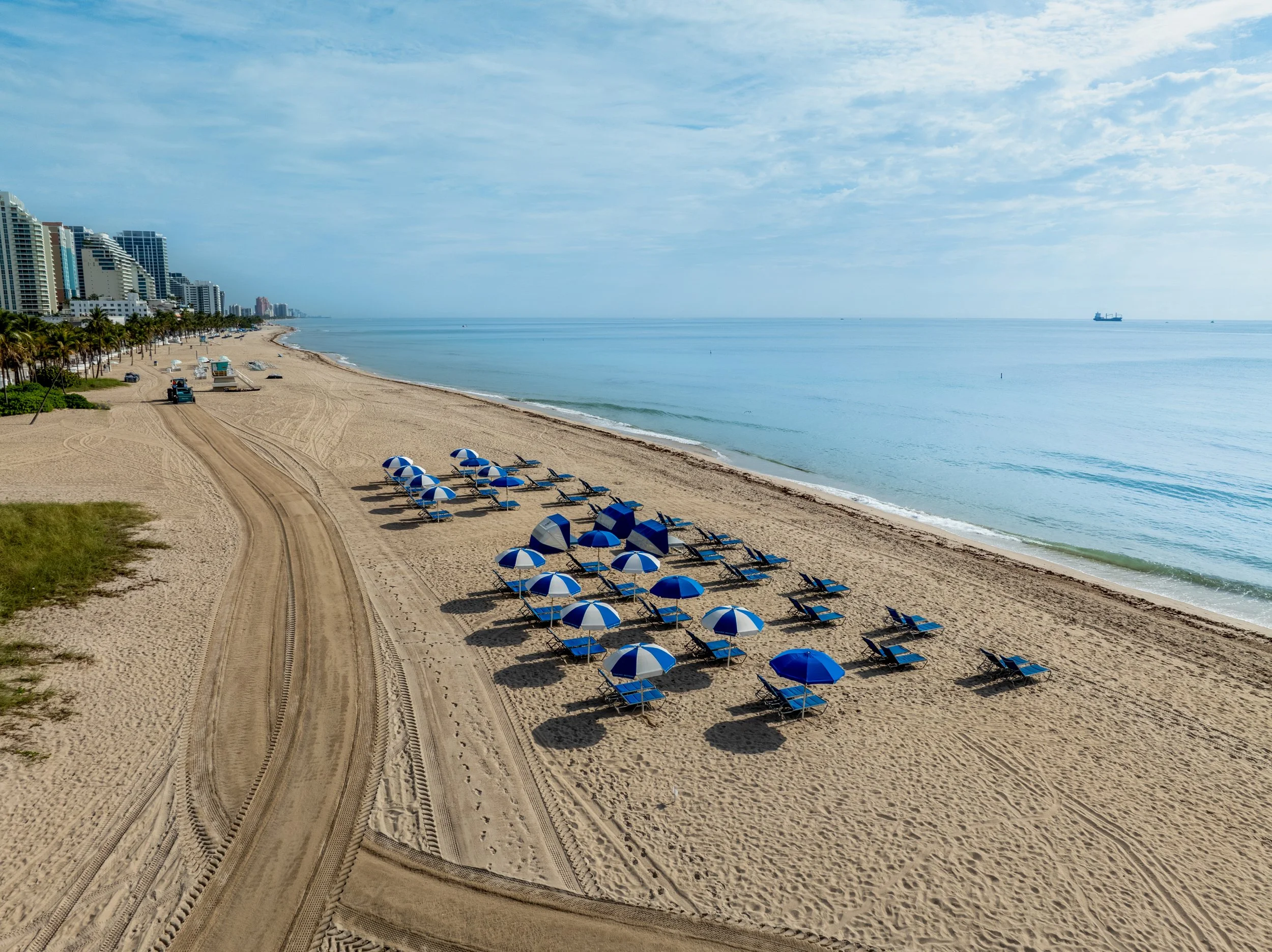 A beach scene with multiple blue and white striped umbrellas and lounge chairs on the sandy shore, as well as high-rise buildings along the coast and a cargo ship in the distance over the calm ocean under a partly cloudy sky.