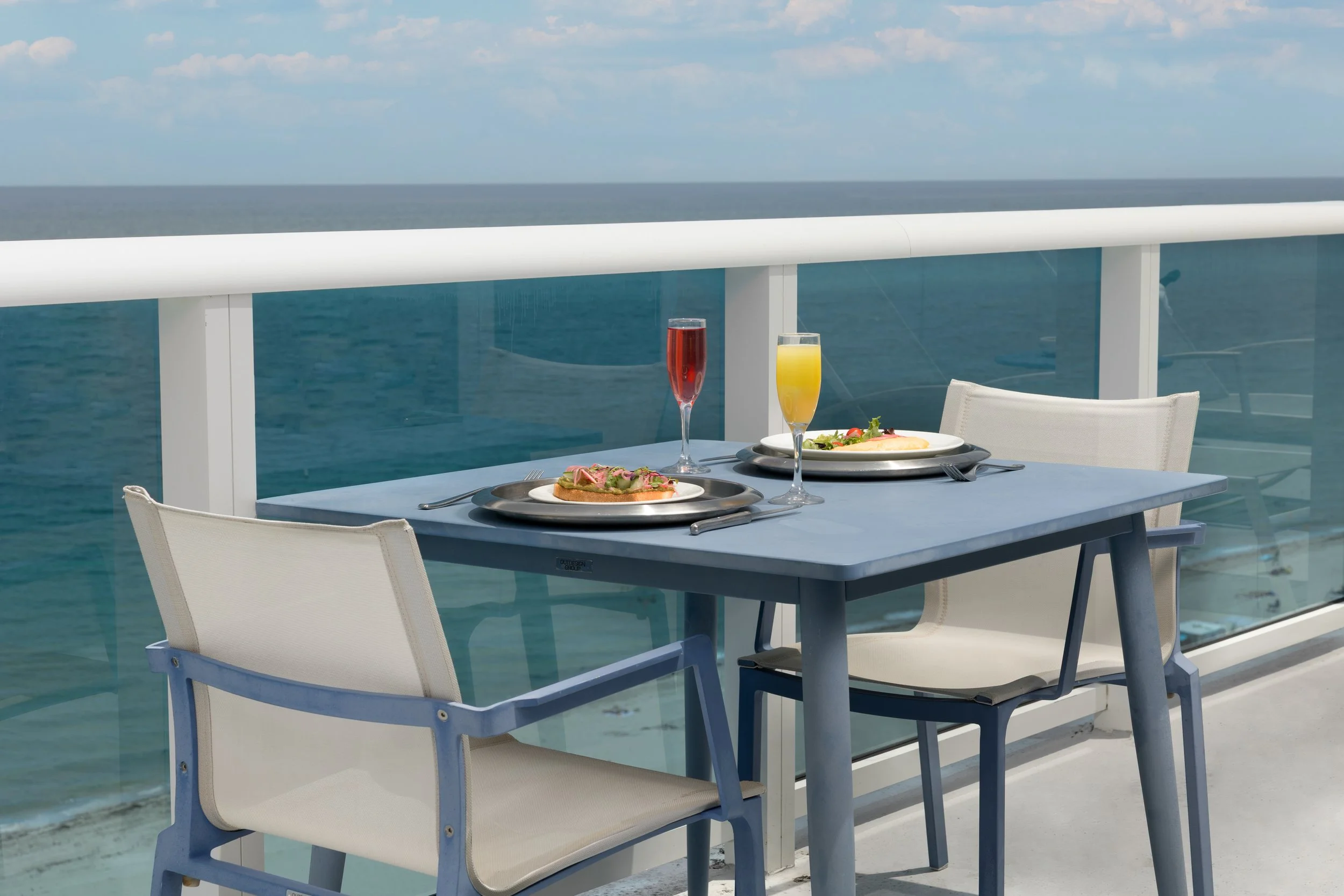 A table set for a meal on a balcony overlooking the ocean. There are two plates with food, two glasses of colorful drinks, and utensils on the table.