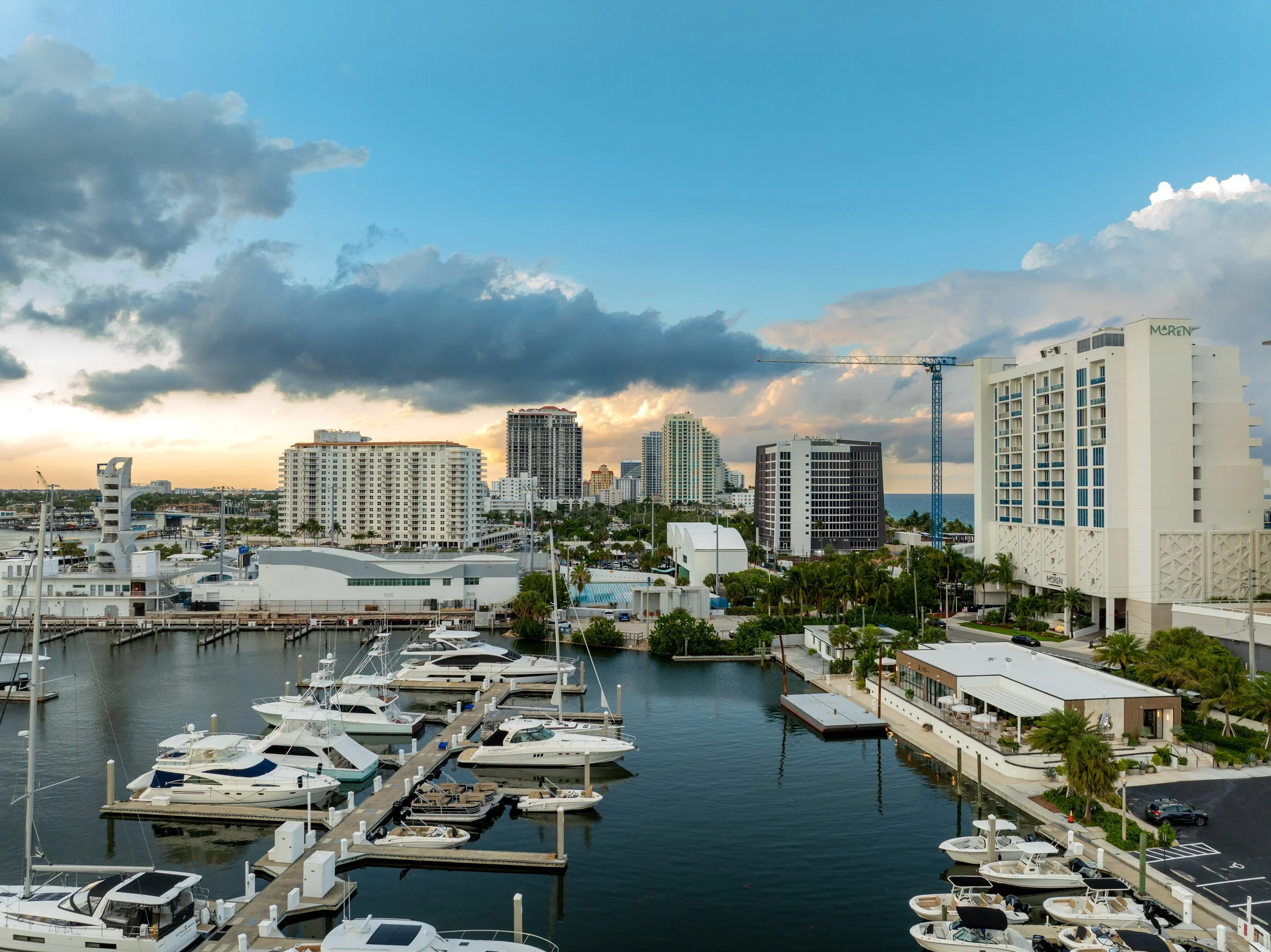 View of a marina with docked boats and yachts, surrounded by high-rise buildings and a city skyline under a partly cloudy sky at sunset.
