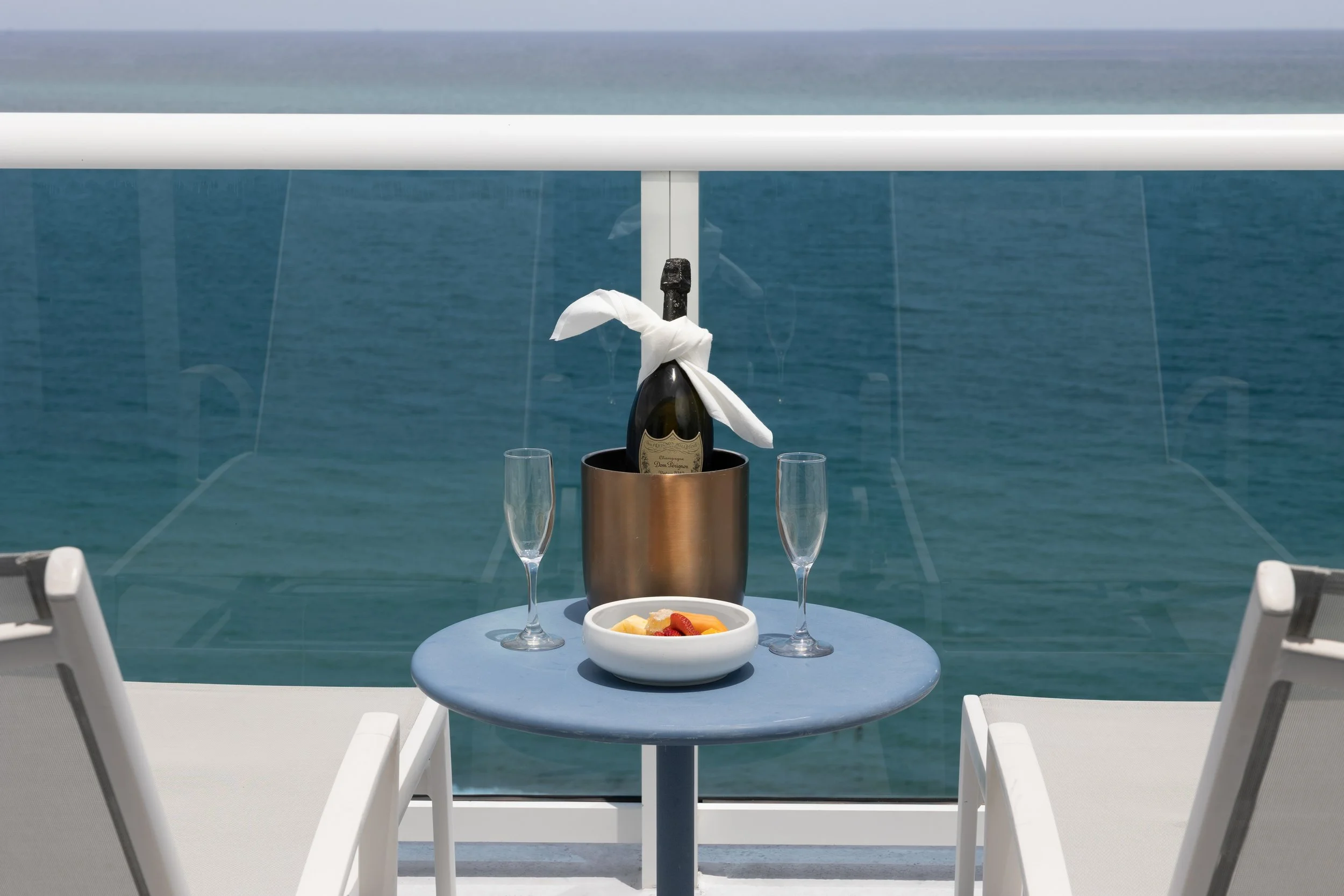 A table set for two with a bottle of champagne in an ice bucket, two champagne glasses, a fruit salad bowl, and a plate of sliced fruit, on a balcony overlooking the ocean.