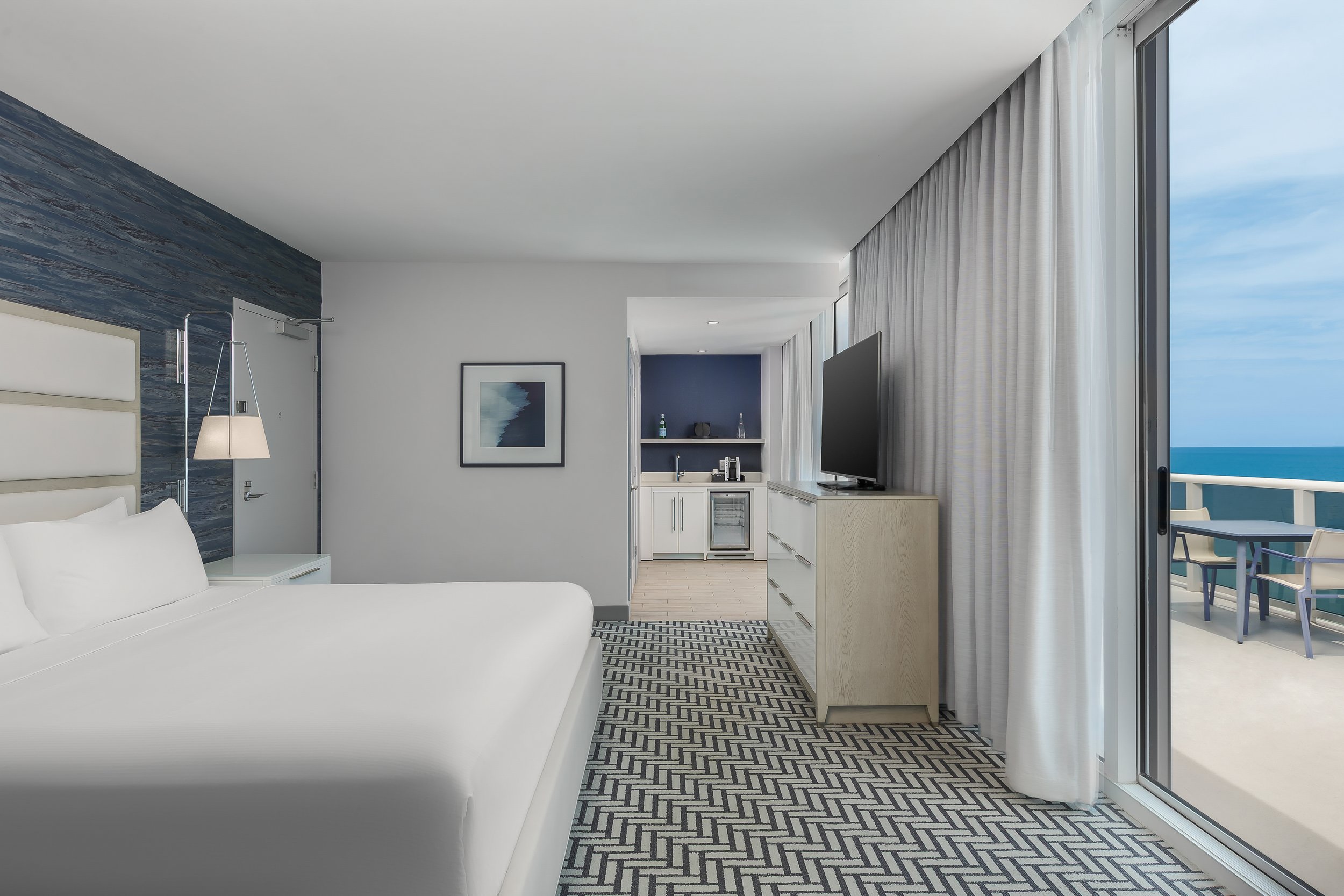 Interior view of a modern hotel room with a white bed, patterned rug, wall art, a small kitchenette, and a large window showing a view of the ocean and balcony.
