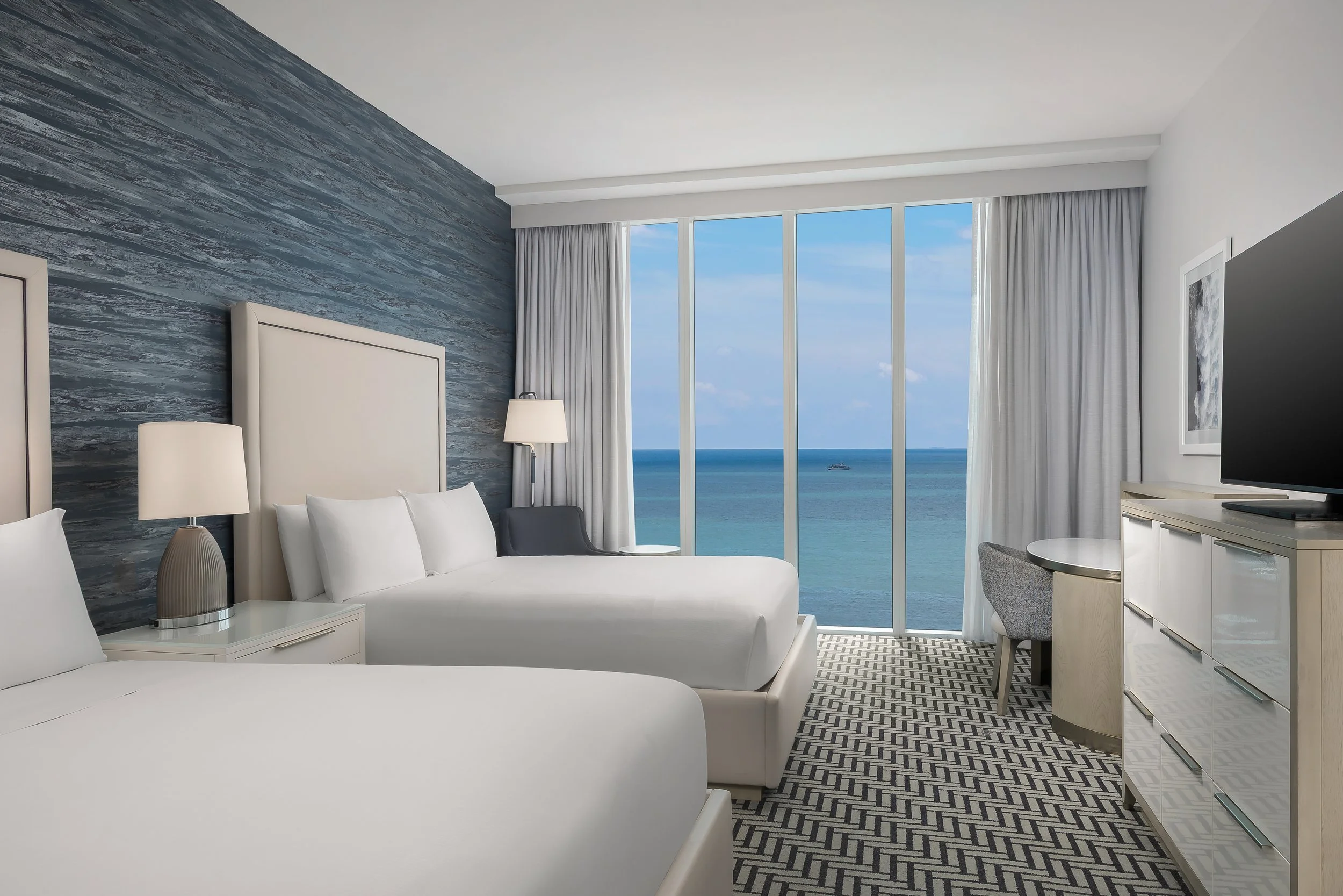 A modern hotel room with a view of the ocean through large windows, featuring a white bed, bedside lamps, a patterned carpet, and minimal decor.