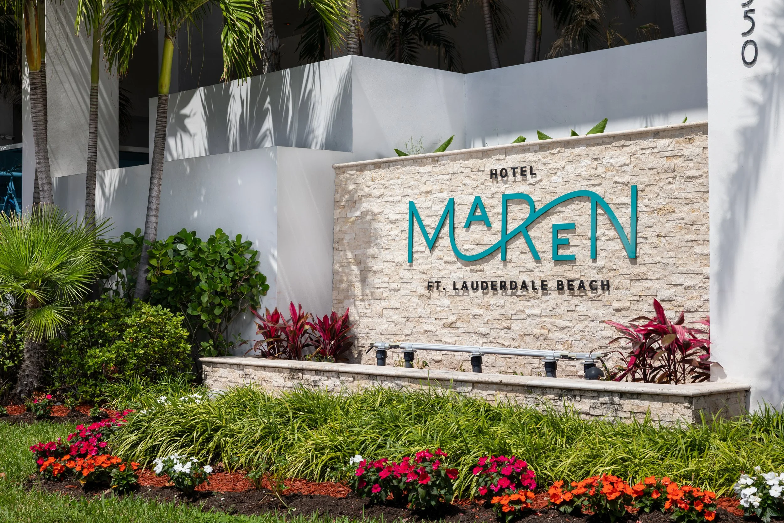 Entrance sign for Hotel Madri at Fort Lauderdale Beach, surrounded by well-maintained colorful flowers and lush green plants, with palm trees and modern white building in the background.